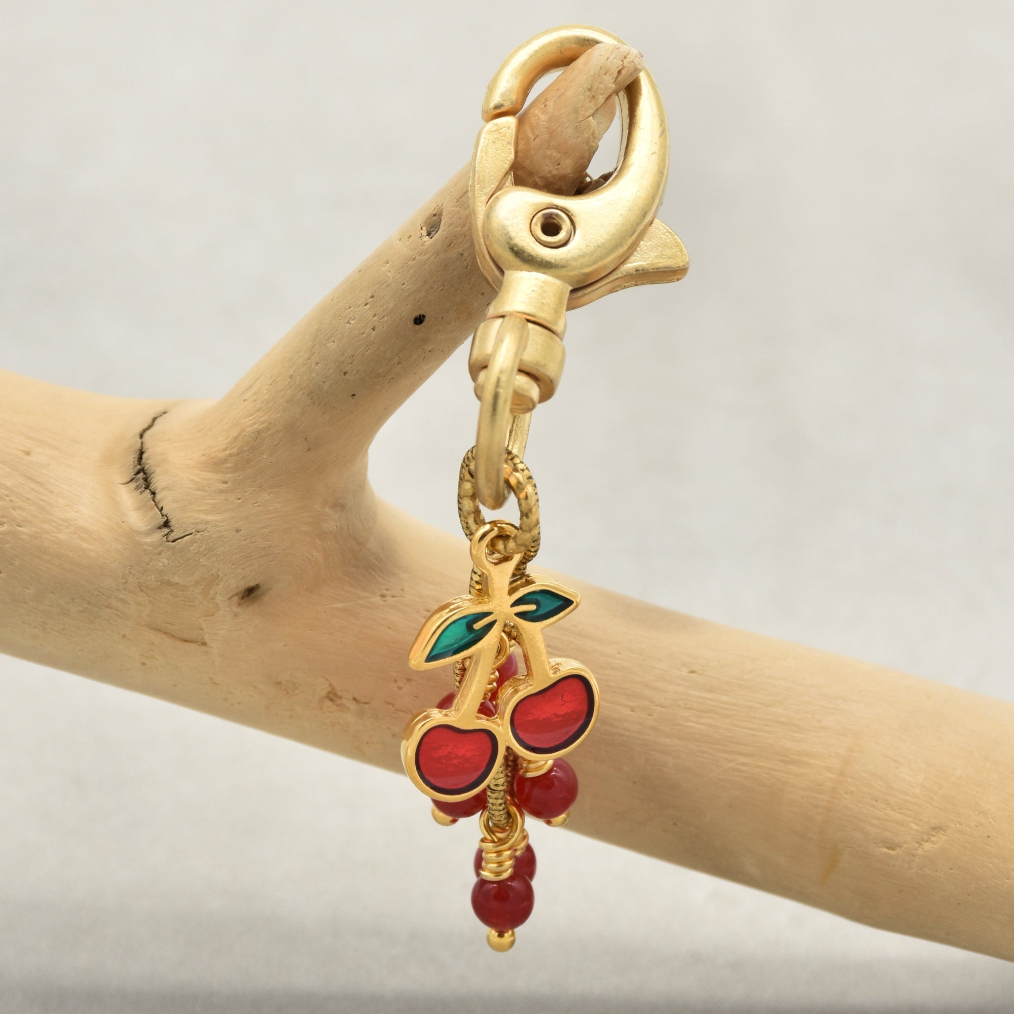 A gold-toned lobster clasp keychain or bag charm featuring three red cherries with green leaves and dangling red beads, displayed on a branch - made with love in Colorado by Ruby Acorn.