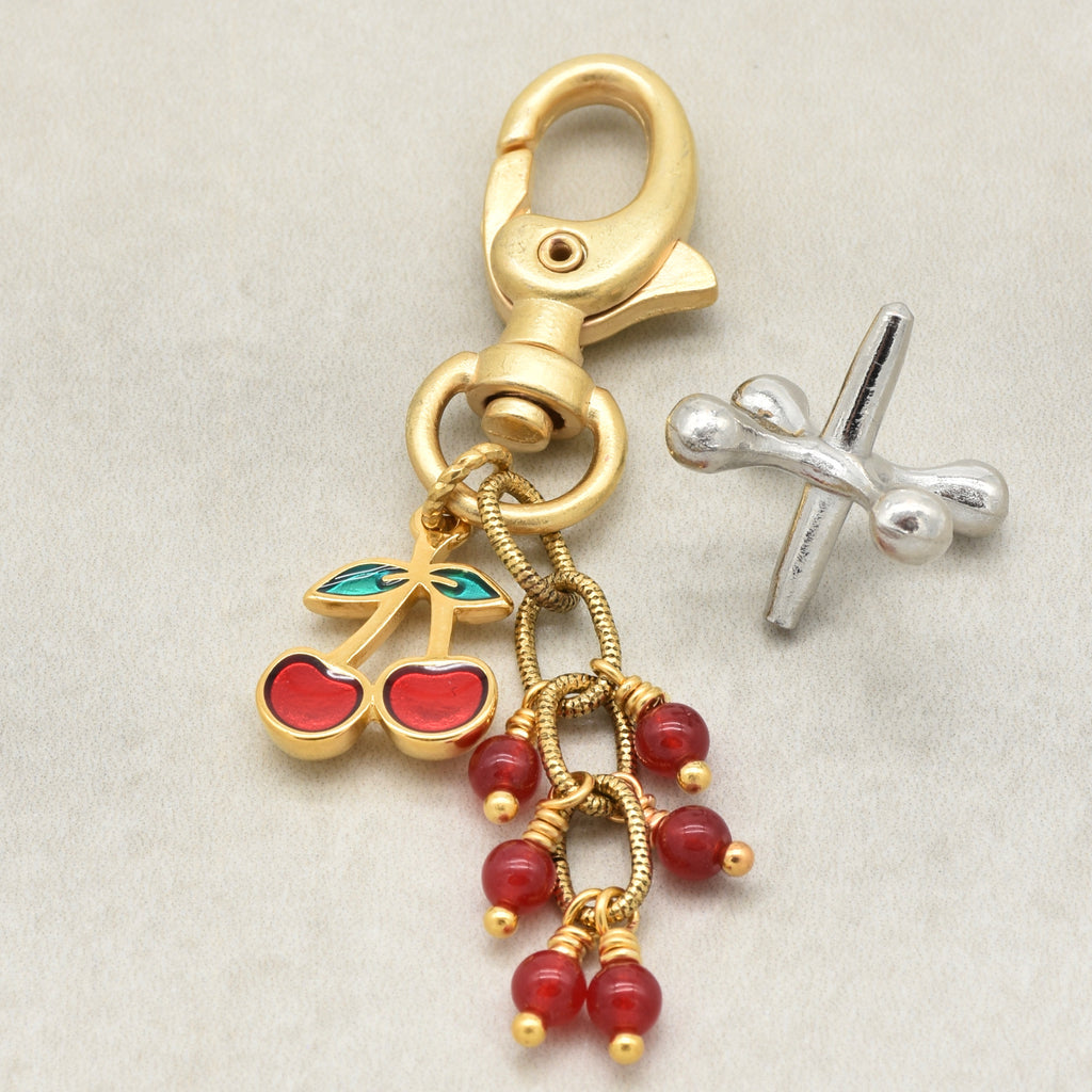 A gold-toned lobster clasp keychain or bag charm with dangling cherry and bead charms in red and green enamel, shown on a light neutral background - made with love in Colorado by Ruby Acorn.
