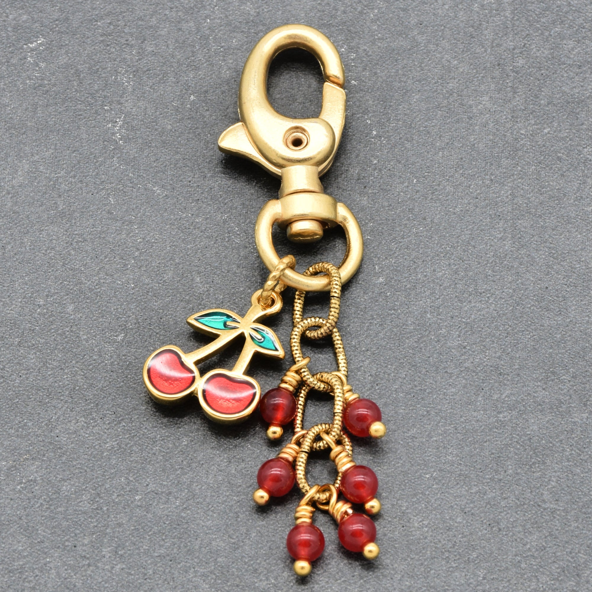 Gold-tone metal keychain charm with a lobster clasp, cherry pendant with green and red enamel, and dangling red bead chains on a dark gray textured background - handmade with love in Colorado by Ruby Acorn.