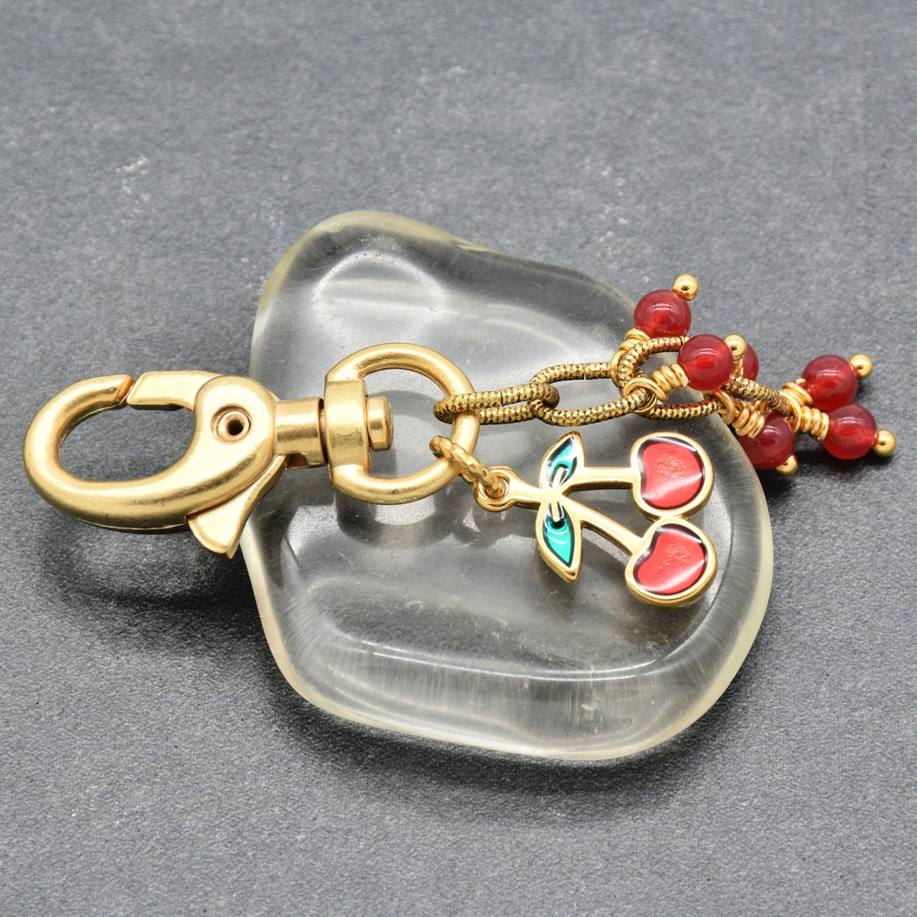 A gold-toned keychain with a lobster clasp, dangling cherry charms with red enamel and green leaves, set against a dark gray textured background - made with love in Colorado by Ruby Acorn.