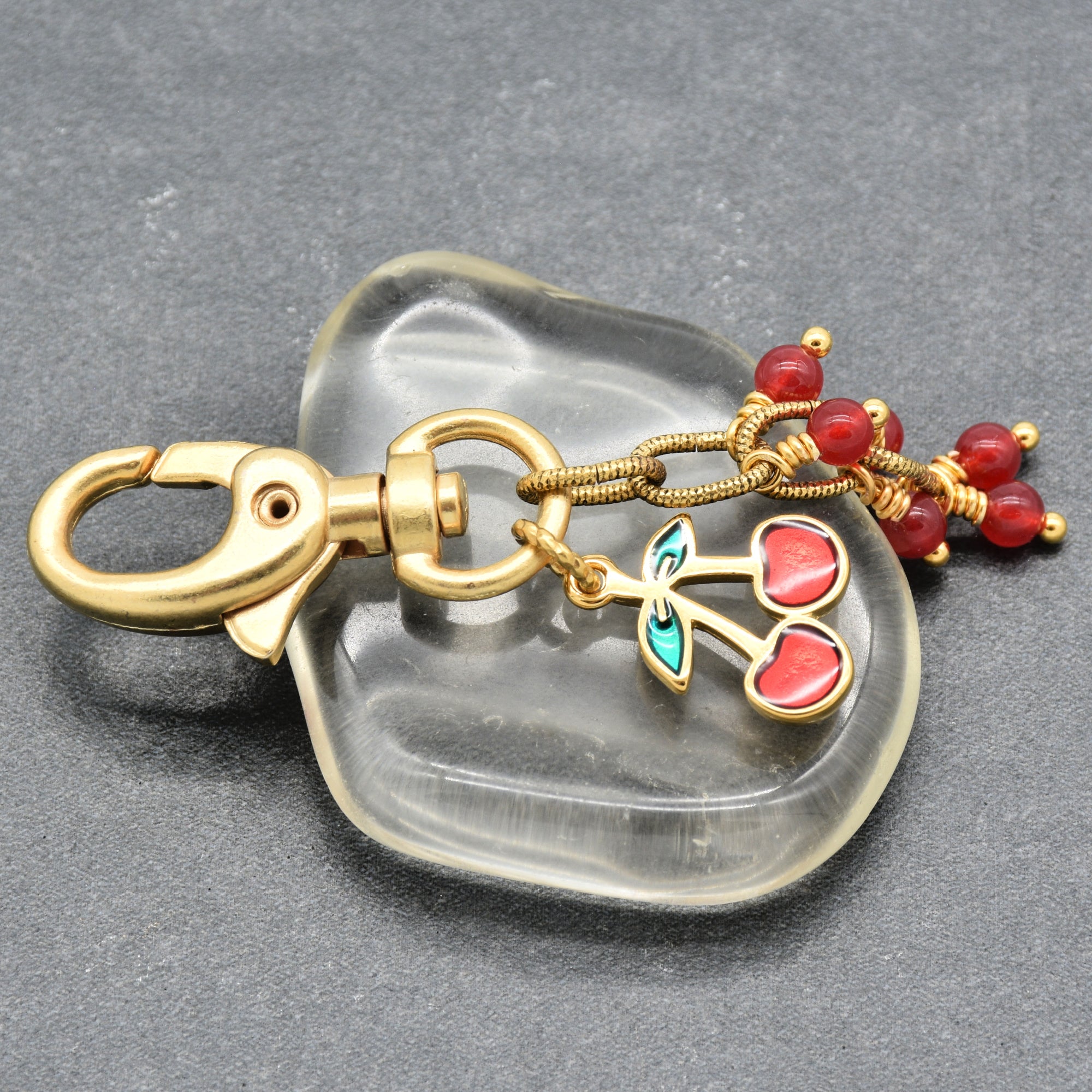 A gold-toned keychain with a lobster clasp, dangling cherry charms with red enamel and green leaves, set against a dark gray textured background - made with love in Colorado by Ruby Acorn.