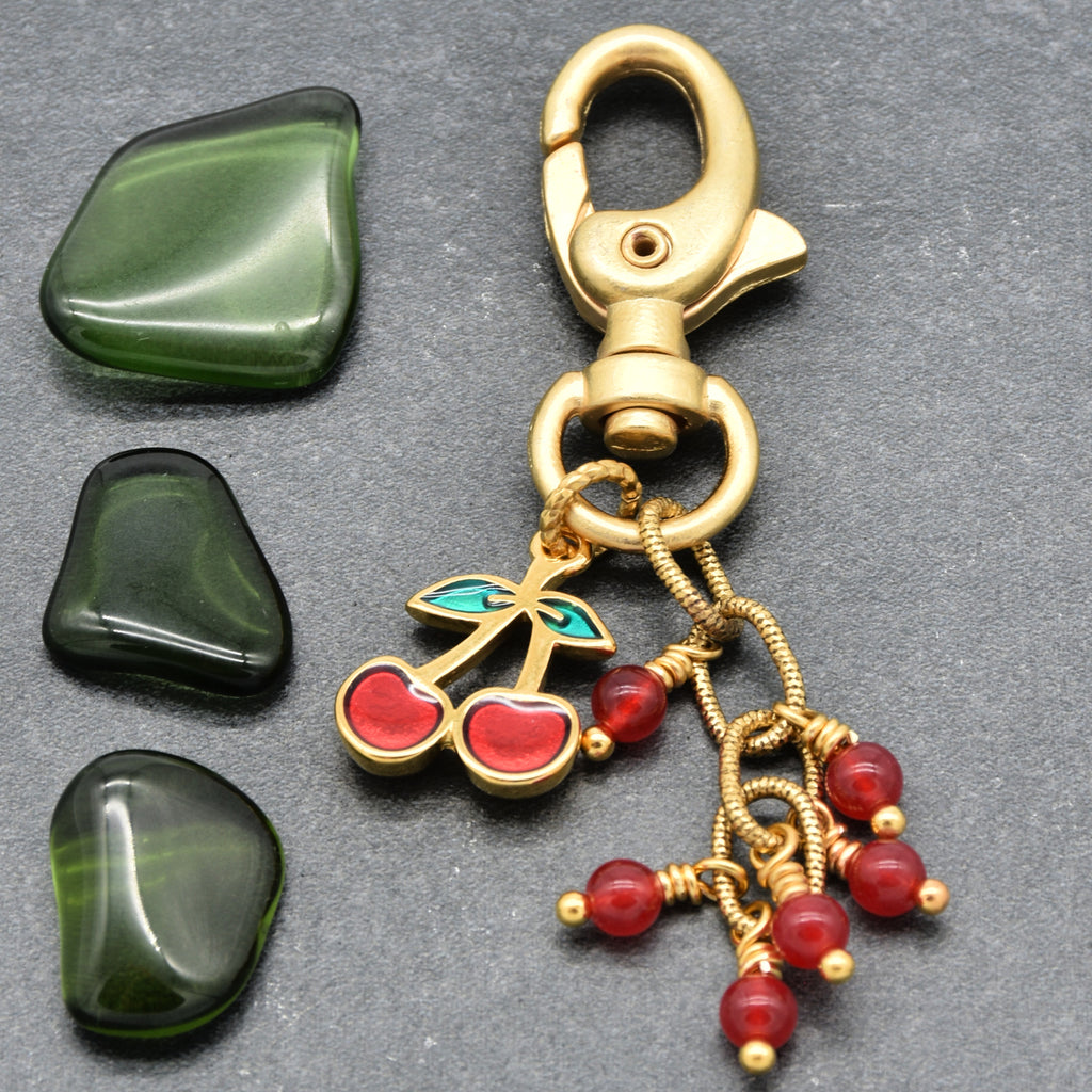 Gold-tone metal keychain with a lobster clasp, a cherry charm with red enamel and green leaf accents, and dangling red jade bead strands, displayed on a dark gray surface with three polished green stones beside it - made with love in Colorado by Ruby Acorn.