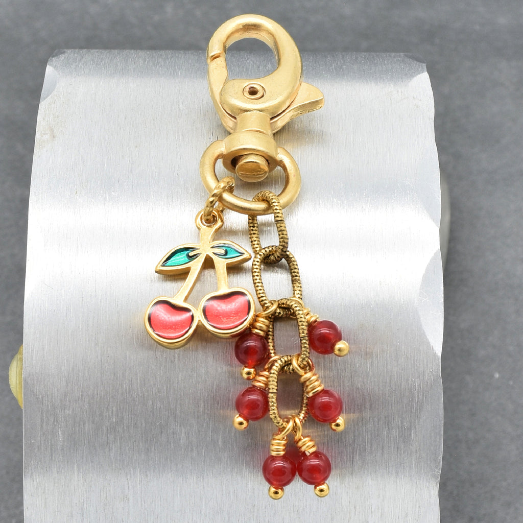A gold-toned lobster clasp keychain or bag charm with dangling cherry and bead charms in red and green enamel, shown on a silver background - made with love in Colorado by Ruby Acorn.