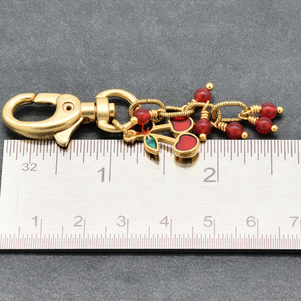 Gold-tone metal keychain with a lobster clasp, red bead accents, and cherry charm, shown next to a metal ruler for scale on a dark gray background - made with love in Colorado by Ruby Acorn.