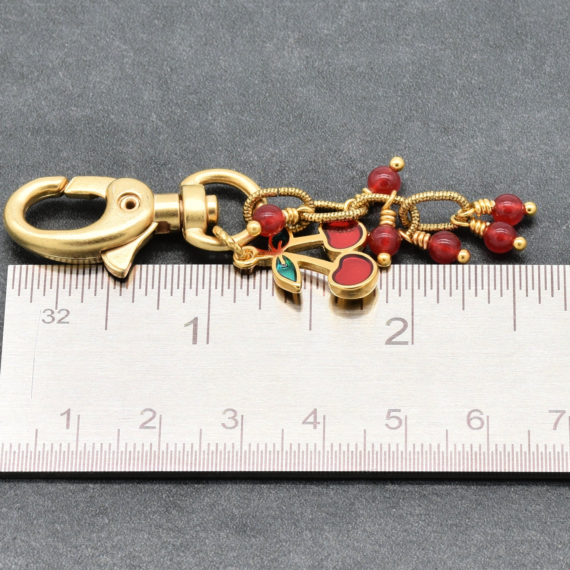 Gold-tone metal keychain with a lobster clasp, red bead accents, and cherry charm, shown next to a metal ruler for scale on a dark gray background - made with love in Colorado by Ruby Acorn.