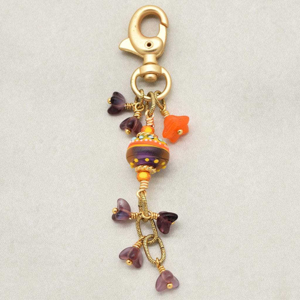 A gold-tone keychain clasp with dangling charms, including a colorful striped glass bead, an orange bellflower bead, and purple flower-shaped beads, shown on a light gray background - made with love in Colorado by Ruby Acorn.