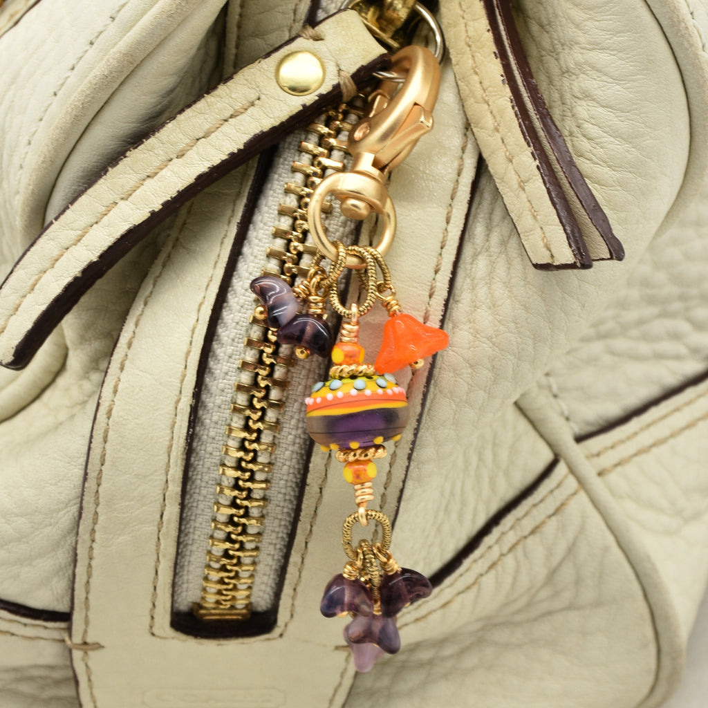 A close-up of a beige textured leather handbag featuring a gold-tone zipper and a decorative multicolor glass charm keychain with purple beads and orange accents - made with love in Colorado by Ruby Acorn.