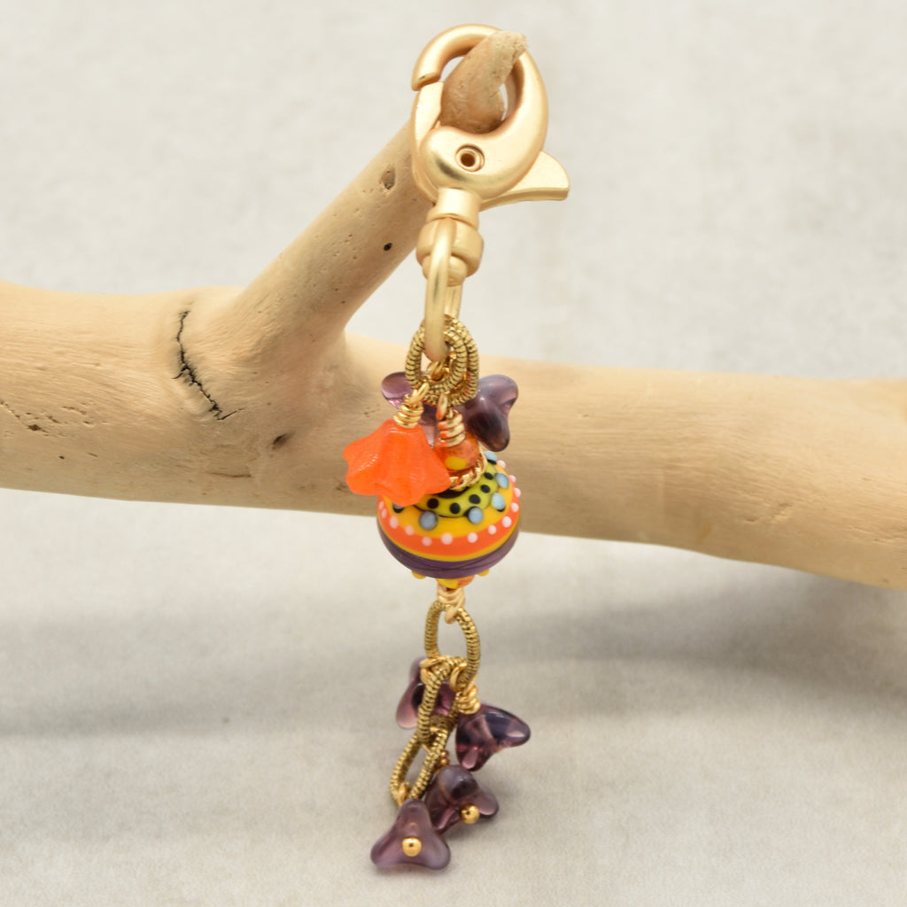 A gold-toned lobster clasp keychain or bag charm with a colorful decorative bead, orange flower, and dangling purple flower beads, displayed on a wooden branch against a light gray background - made with love in Colorado by Ruby Acorn.