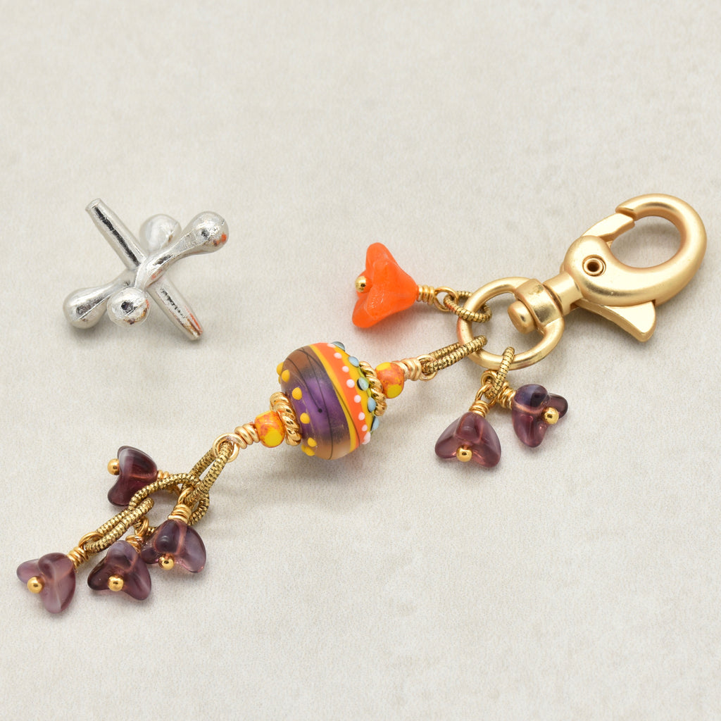 A gold-toned keychain charm with a large decorative multicolor glass bead, small orange beads, and purple flower-shaped beads, displayed on a light beige background - made with love in Colorado by Ruby Acorn.