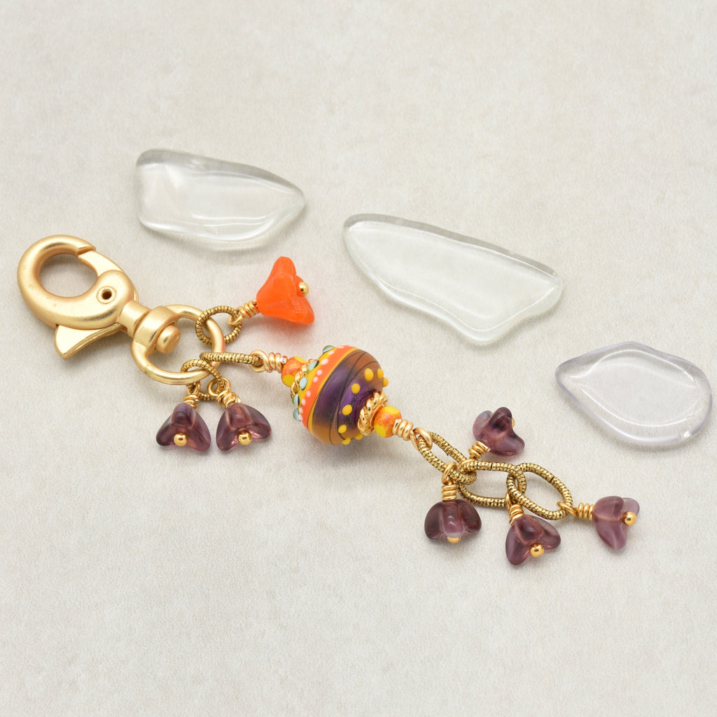 A handmade glass bead keychain charm with a gold clasp, featuring a central multicolor striped bead, and small purple glass flower charms, on a light beige background - made with love in Colorado by Ruby Acorn.