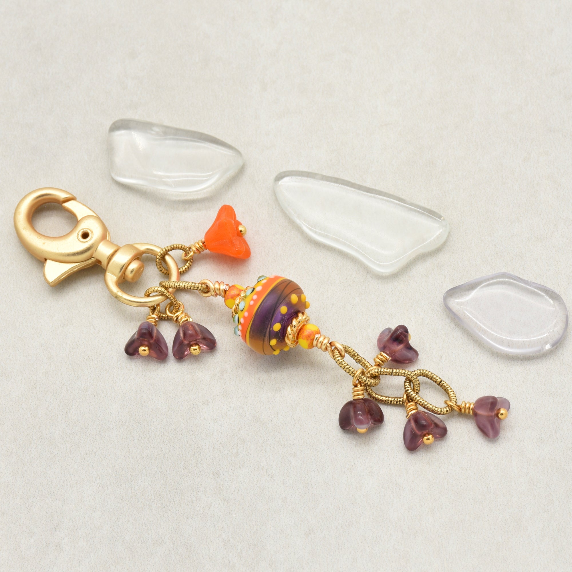 A handmade glass bead keychain charm with a gold clasp, featuring a central multicolor striped bead, and small purple glass flower charms, on a light beige background - made with love in Colorado by Ruby Acorn.