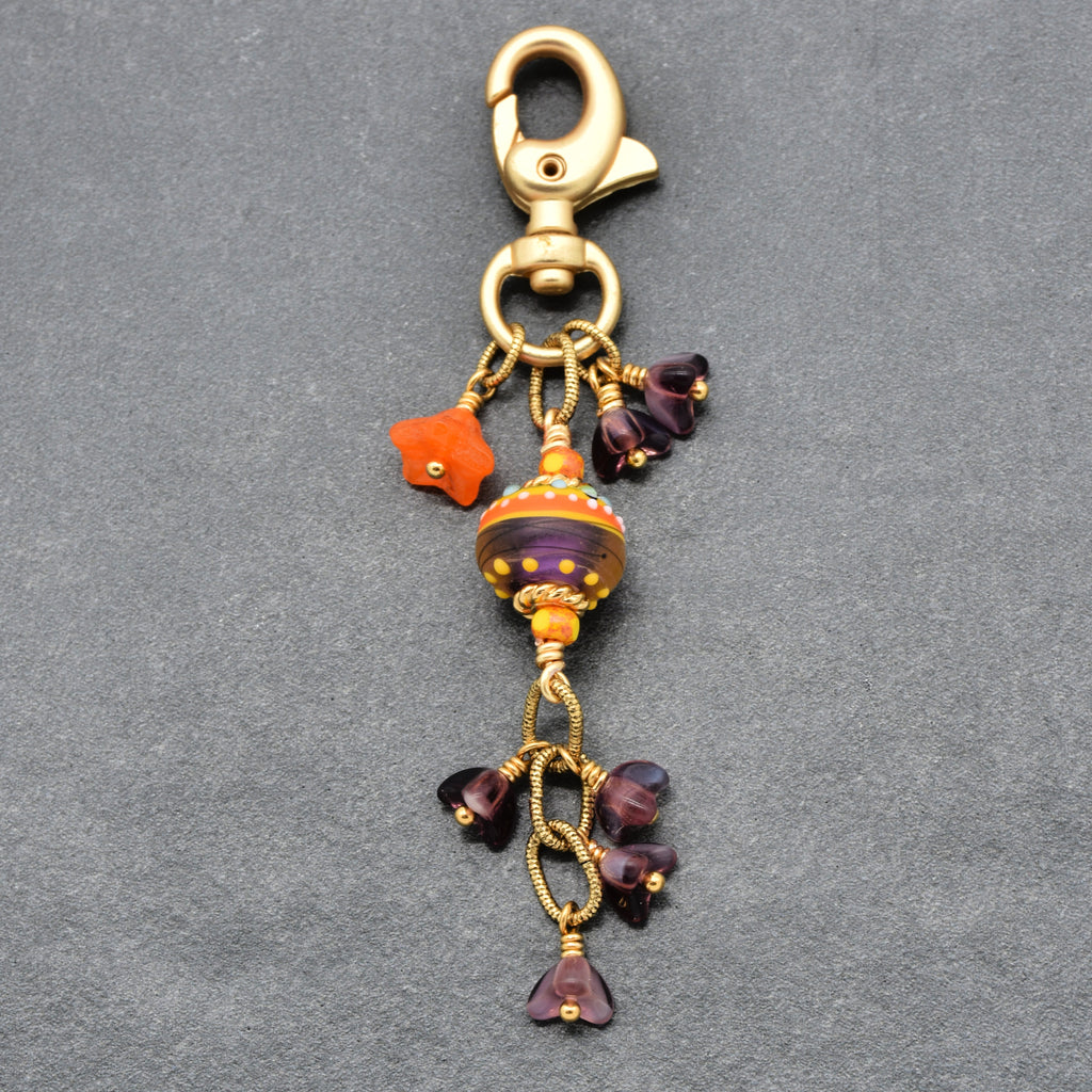 A decorative lobster clasp keychain featuring a central striped glass ornament in orange, purple, and gold, surrounded by small purple glass beads and a bright orange star charm, all set against a textured gray background - made with love in Colorado by Ruby Acorn.