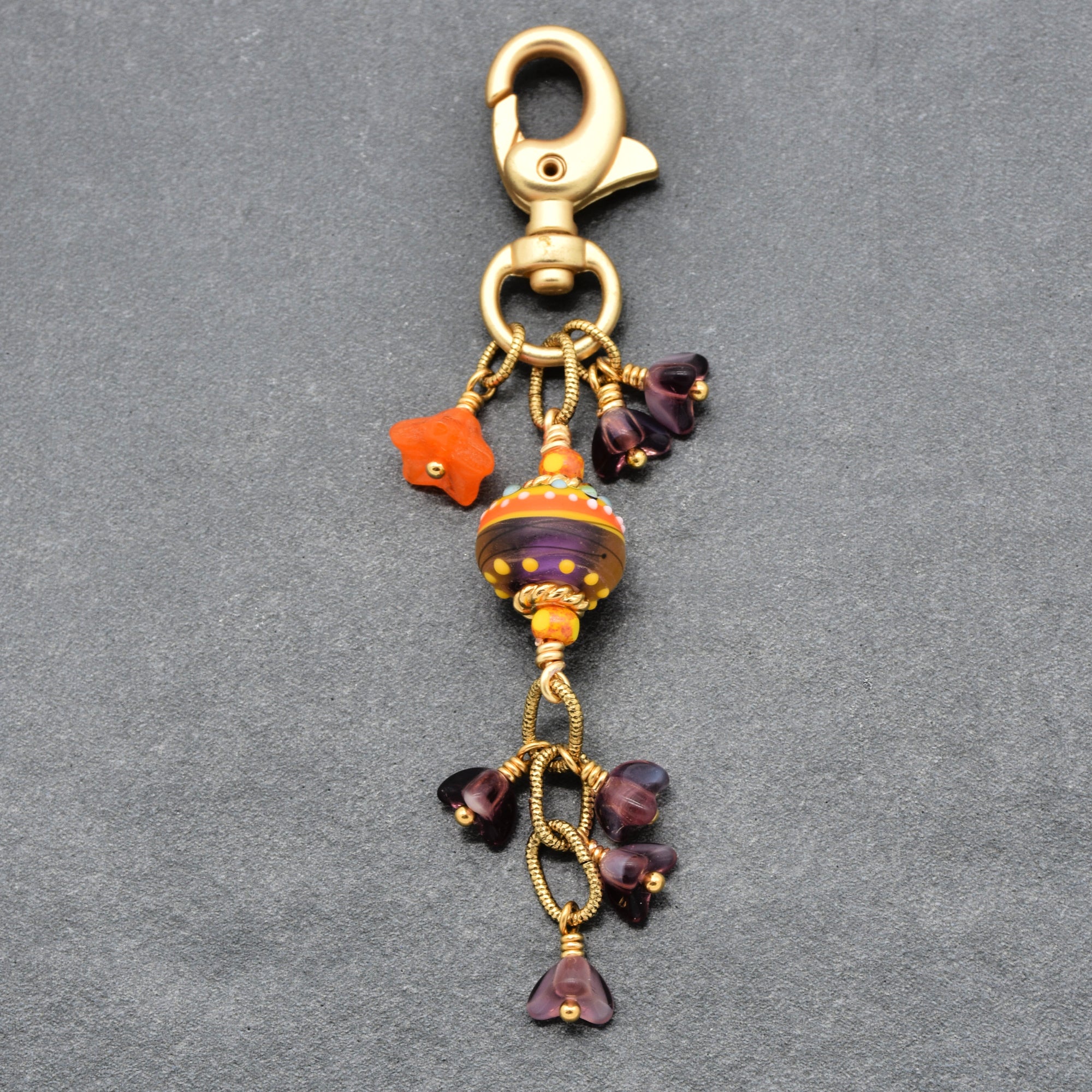 A decorative lobster clasp keychain featuring a central striped glass ornament in orange, purple, and gold, surrounded by small purple glass beads and a bright orange star charm, all set against a textured gray background - made with love in Colorado by Ruby Acorn.