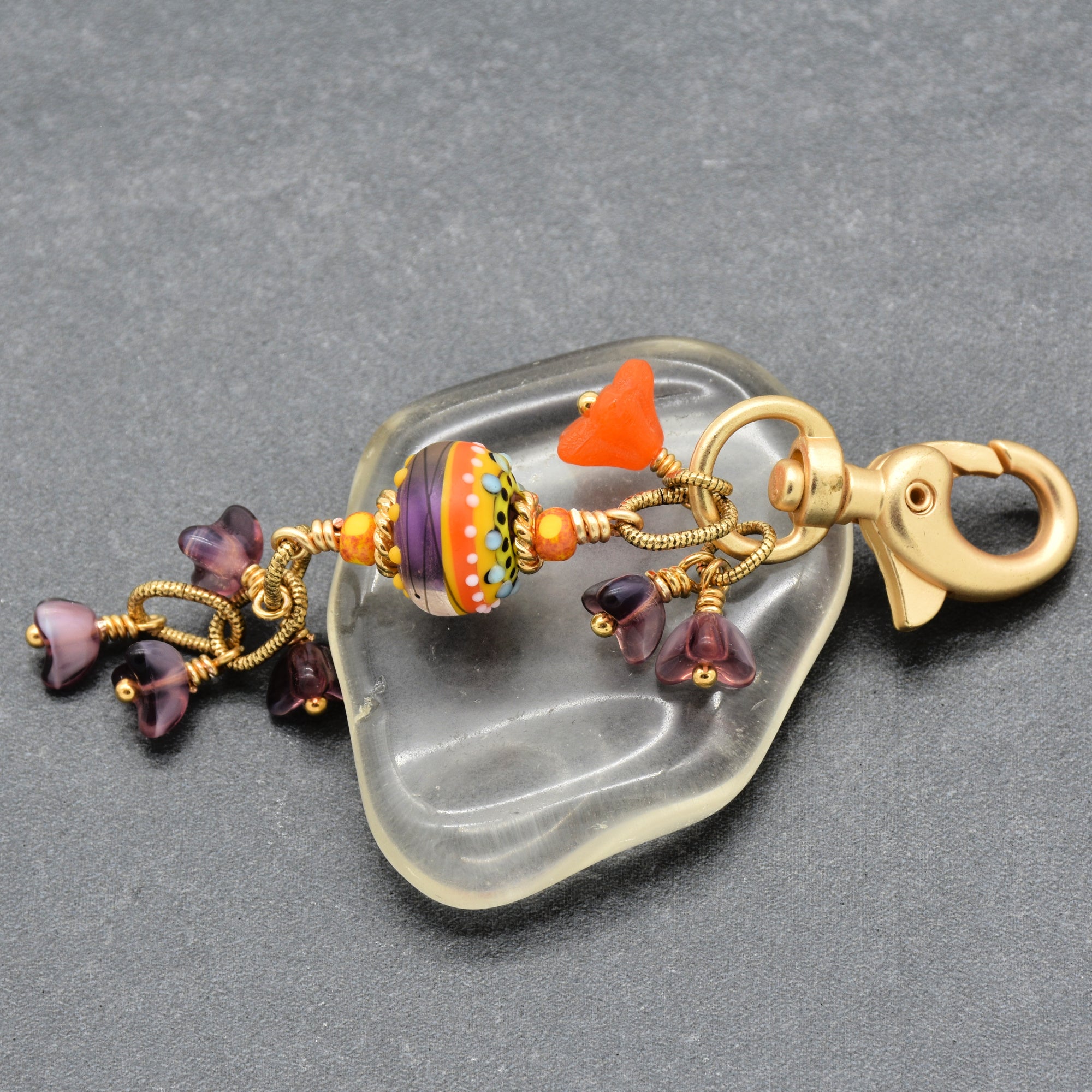 A bohemian-style glass charm keychain with a large patterned orange, yellow, and purple glass bead, small orange beads, purple glass flowers, and a gold lobster clasp, displayed on a clear glass dish over a dark gray background - made with love in Colorado by Ruby Acorn.
