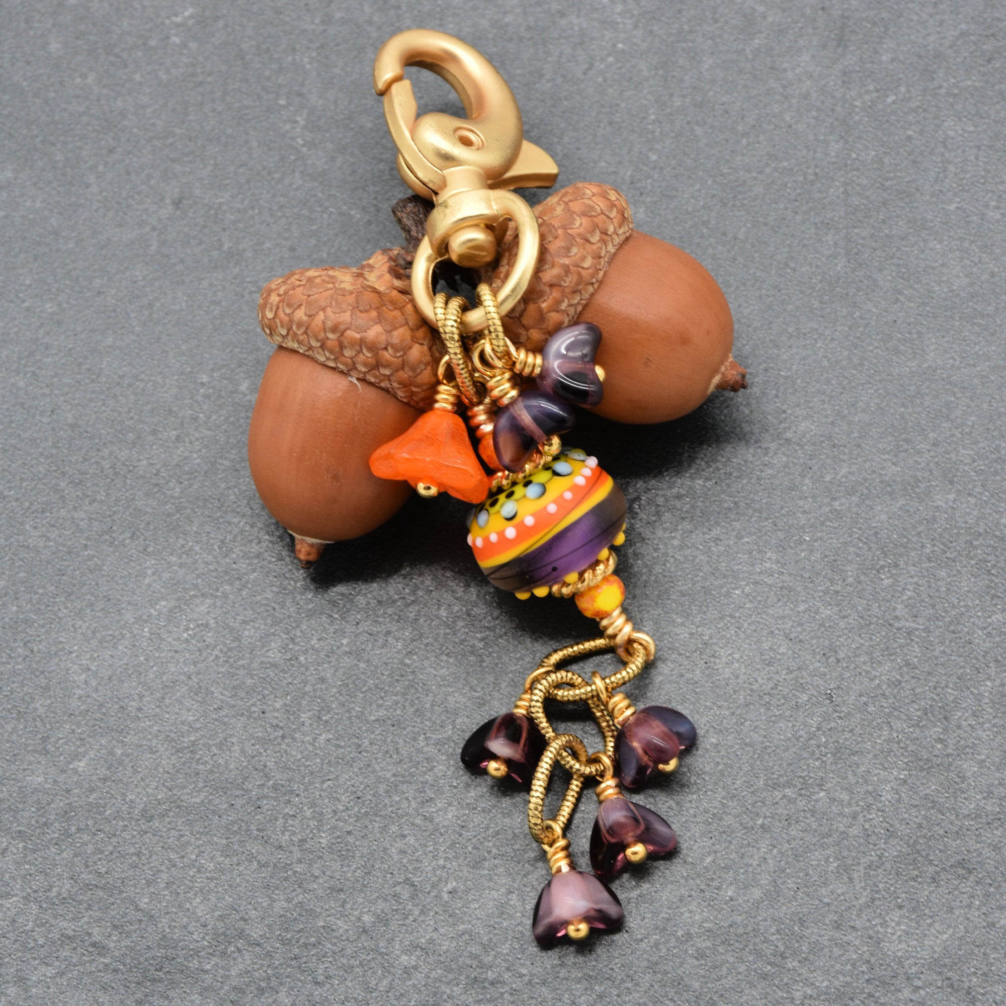 A decorative keychain charm with a gold-tone lobster clasp, a colorful glass bead, and dangling purple crystal flowers, photographed on a textured gray background - made with love in Colorado by Ruby Acorn.