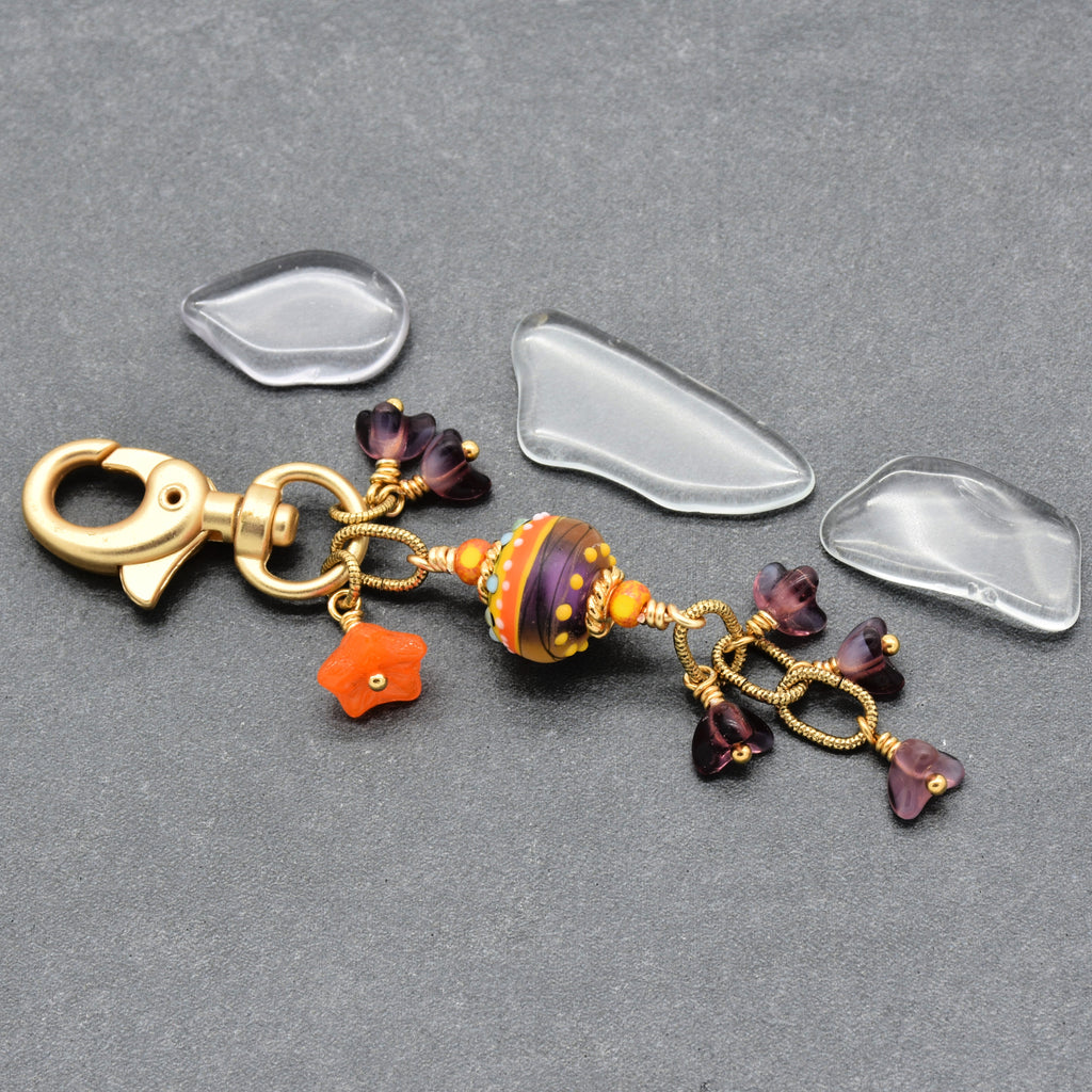 A gold-tone lobster clasp keychain charm with a patterned purple and orange glass bead, small purple flower charms, and an orange star charm, displayed on a gray textured background - made with love in Colorado by Ruby Acorn.