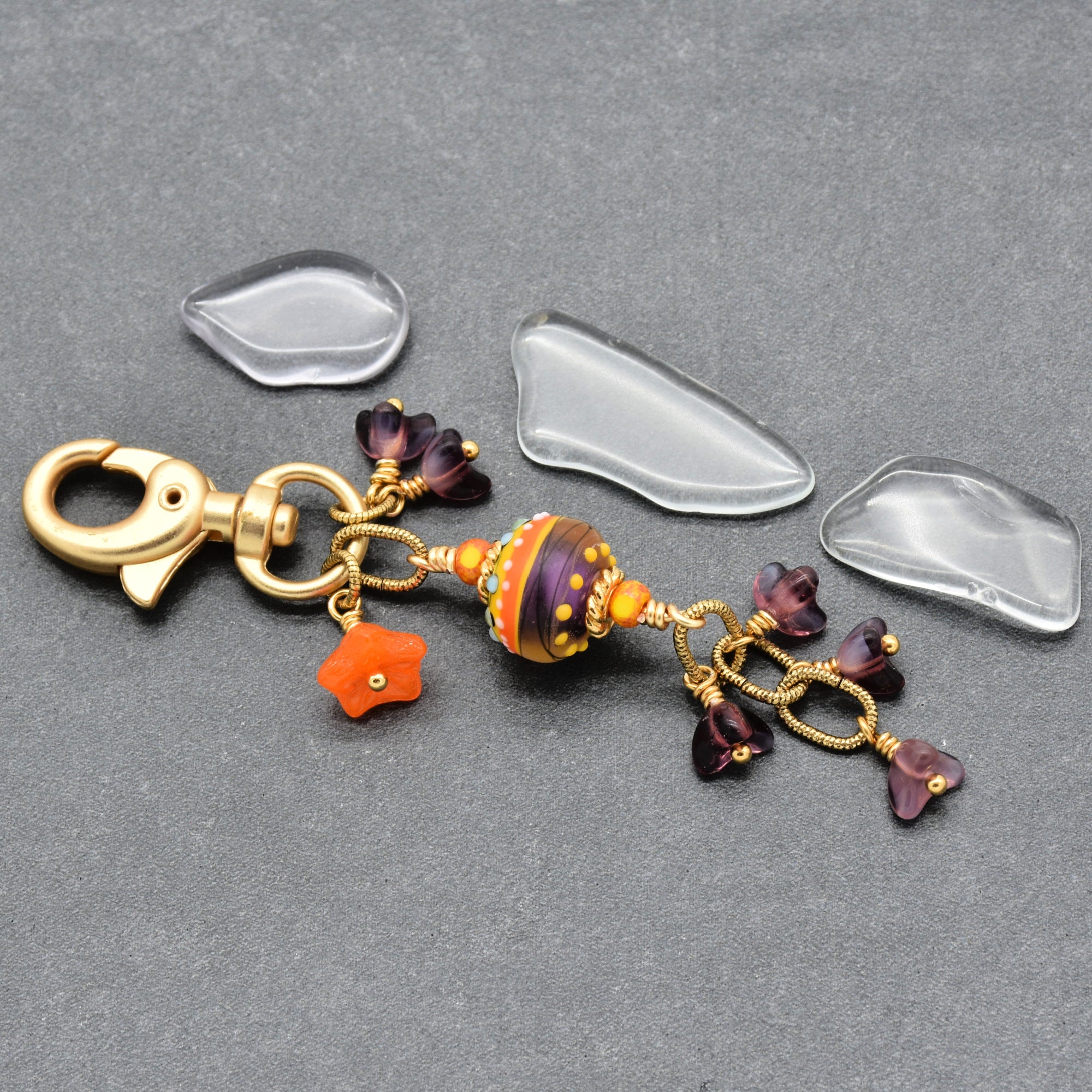 A gold-tone lobster clasp keychain charm with a patterned purple and orange glass bead, small purple flower charms, and an orange star charm, displayed on a gray textured background - made with love in Colorado by Ruby Acorn.