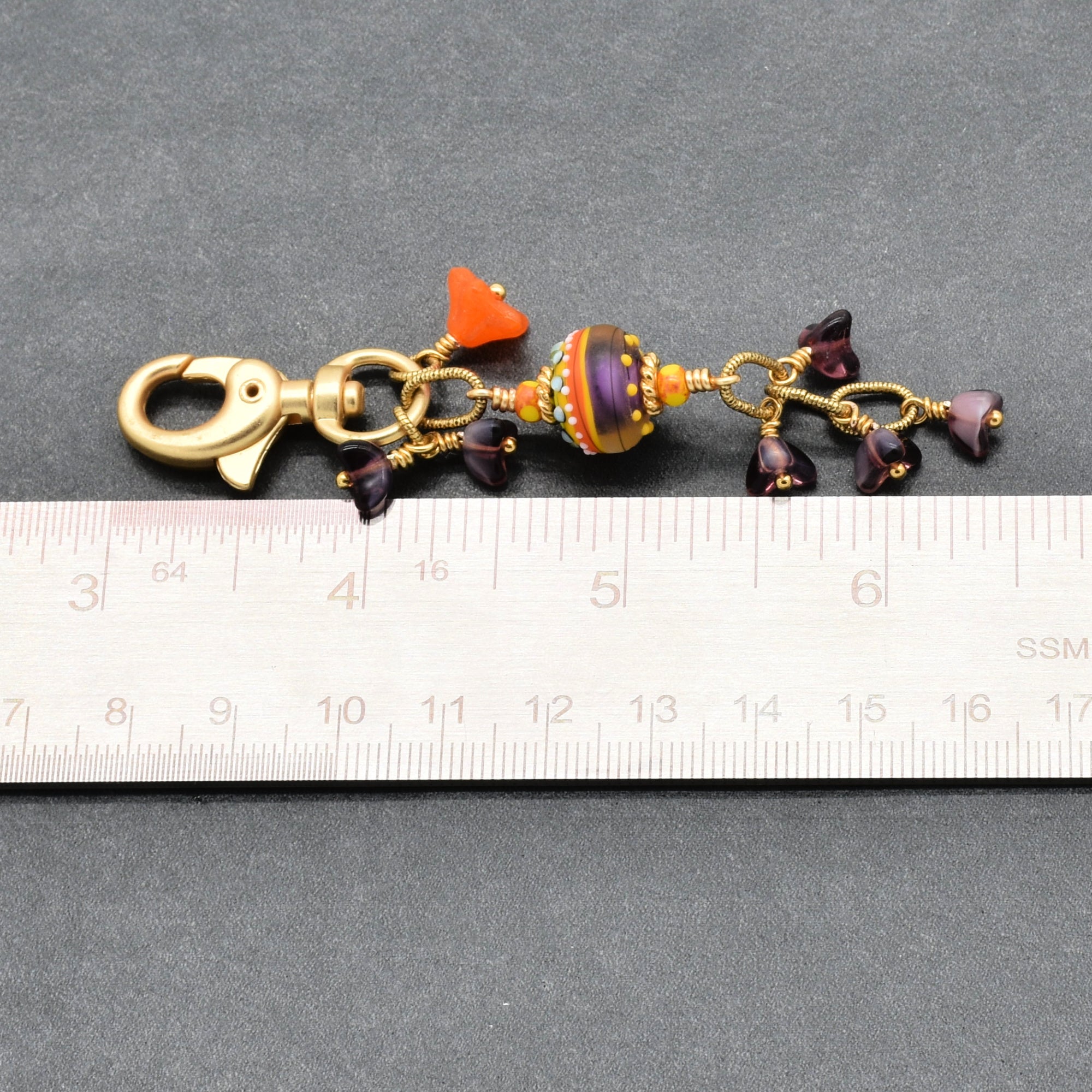 A handmade glass bead keychain with a gold clasp, shown next to a metal ruler for scale on a dark gray background - made with love in Colorado by Ruby Acorn.