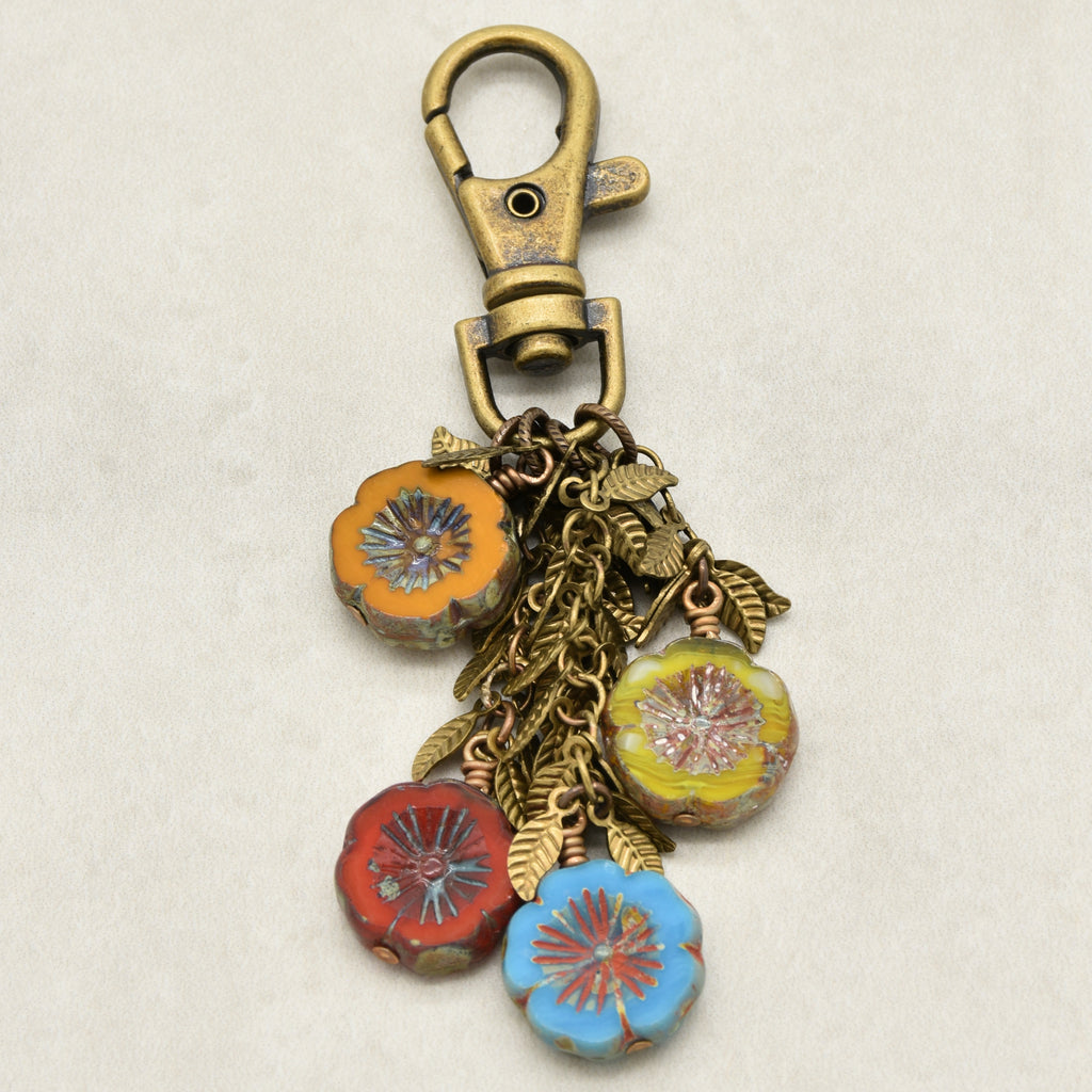 An antique bronze-toned lobster clasp keychain featuring four dangling flower charms in orange, red, yellow, and blue, accented with leaf chain on a light beige background - made with love in Colorado by Ruby Acorn.