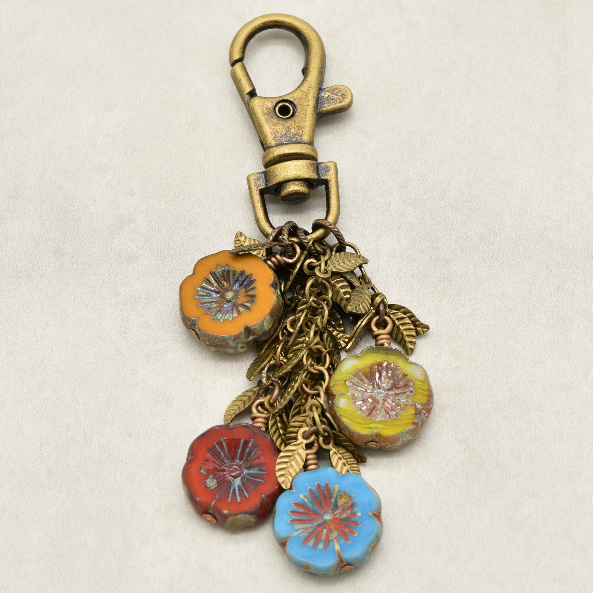 An antique bronze-toned lobster clasp keychain featuring four dangling flower charms in orange, red, yellow, and blue, accented with leaf chain on a light beige background - made with love in Colorado by Ruby Acorn.