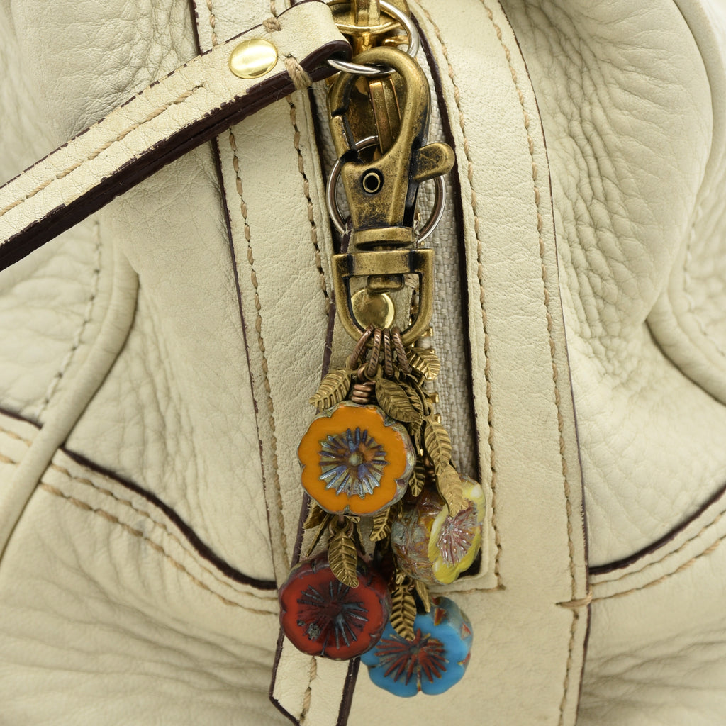 Close-up of a cream-colored leather handbag featuring a brass-tone zipper pull adorned with colorful floral charms in orange, red, blue, and green - made with love in Colorado by Ruby Acorn.