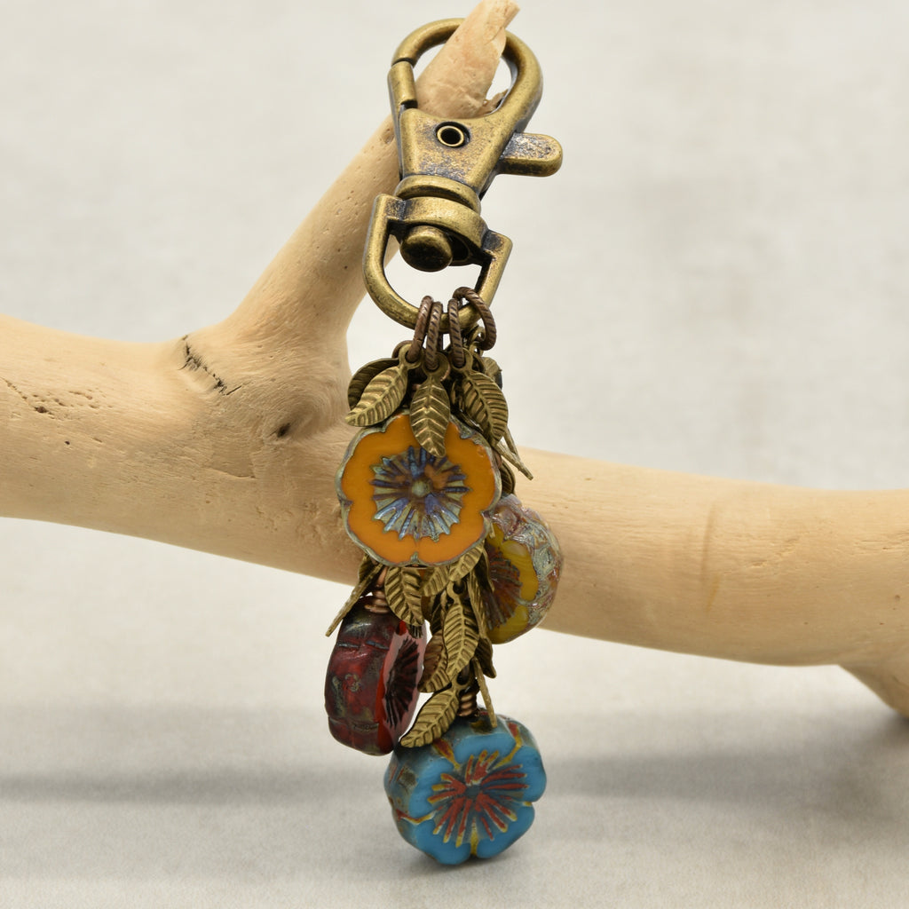 An antique bronze keychain with floral and leaf charms, hanging from a wooden branch. The keychain features a lobster clasp, antique brass-toned metal leaves, and colorful glass beads in orange, green, blue, and red - made with love in Colorado by Ruby Acorn.