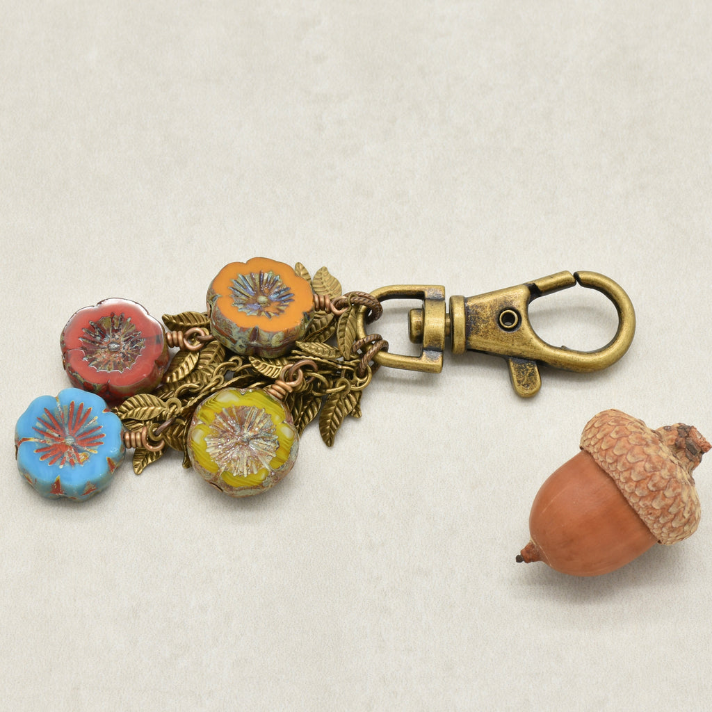 A vintage-style brass keychain or bag charm with four colorful floral beads in blue, red, orange and yellow, accented by antique brass-toned leaf chain, shown next to a decorative acorn on a light beige background - made with love in Colorado by Ruby Acorn.
