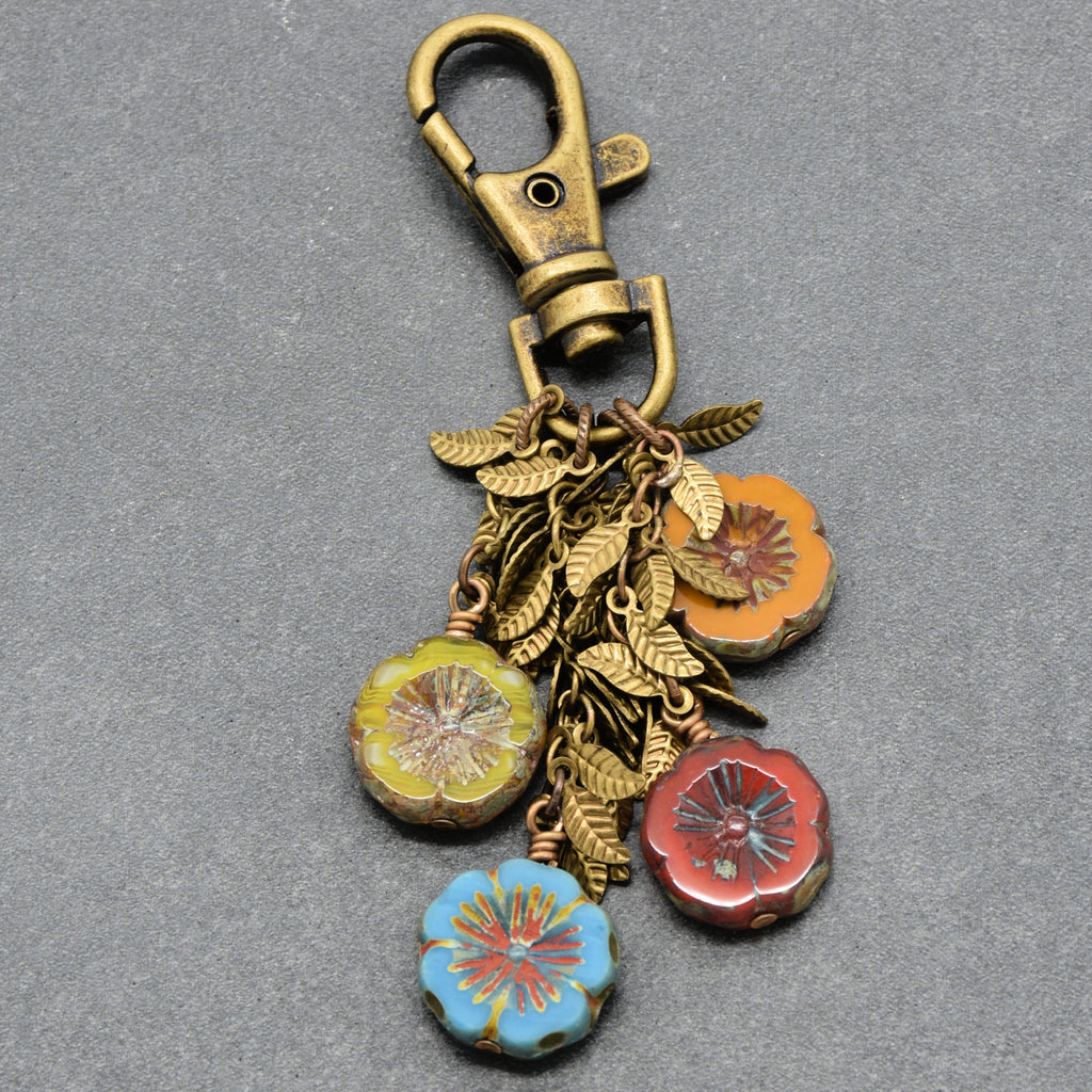 An antique bronze keychain clasp with hanging charms, including gold-toned leaf chains and four colorful glass flower beads in yellow-green, orange, red, and light blue, set against a textured gray background - made with love in Colorado by Ruby Acorn.