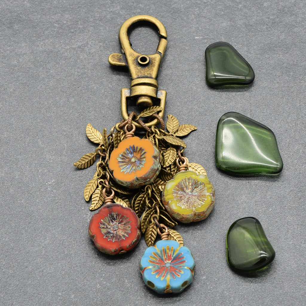 An antique bronze keychain featuring a lobster clasp and four colorful floral glass beads (orange, green, red, and light blue) with antique brass-toned leaf chain, displayed next to three polished green stones on a textured gray background - made with love in Colorado by Ruby Acorn.