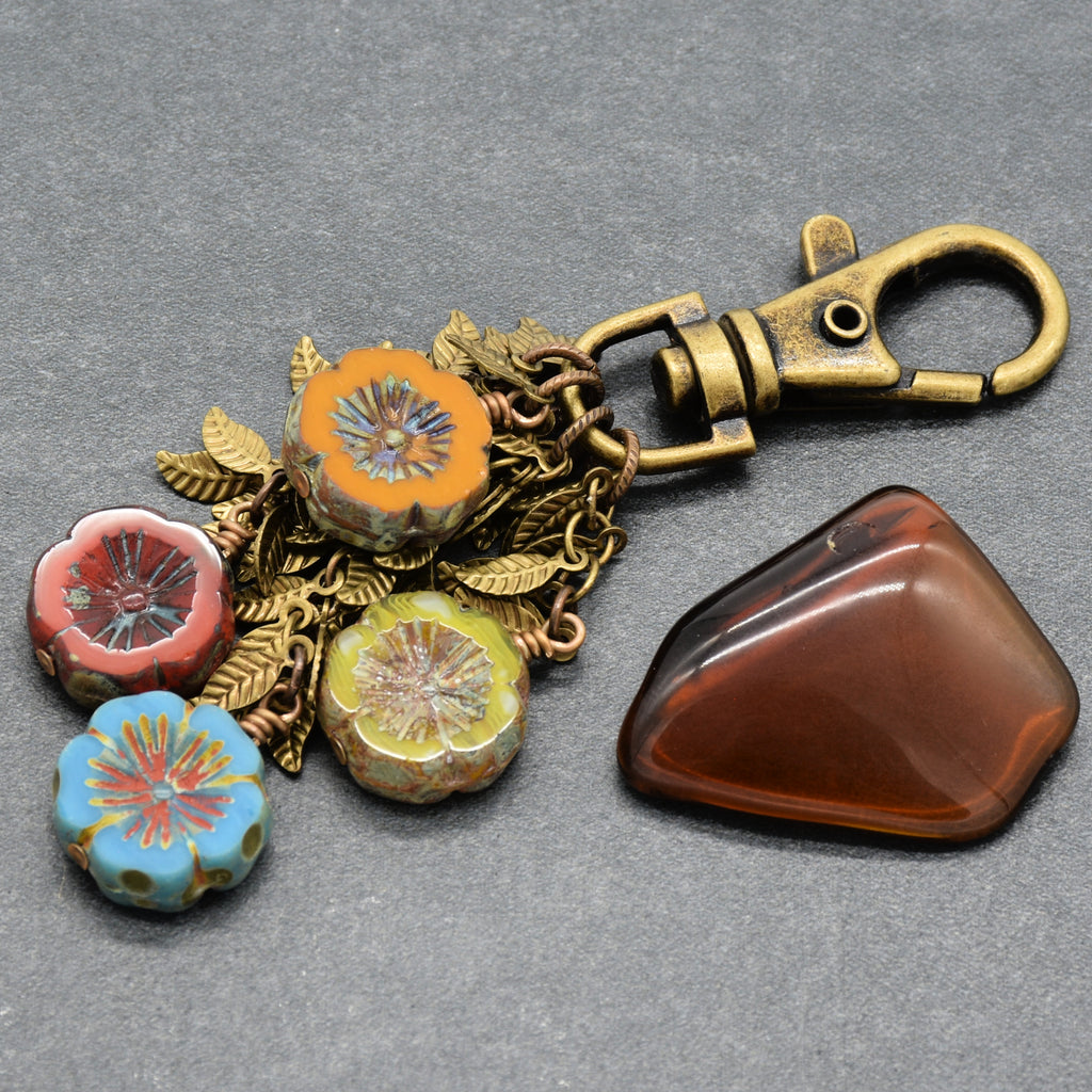A vintage-style metal keychain or bag charm with a brass-toned clasp, floral bead accents in orange, pink, yellow, and blue, and antique brass falling leaves chain, on a gray background - made with love in Colorado by Ruby Acorn.