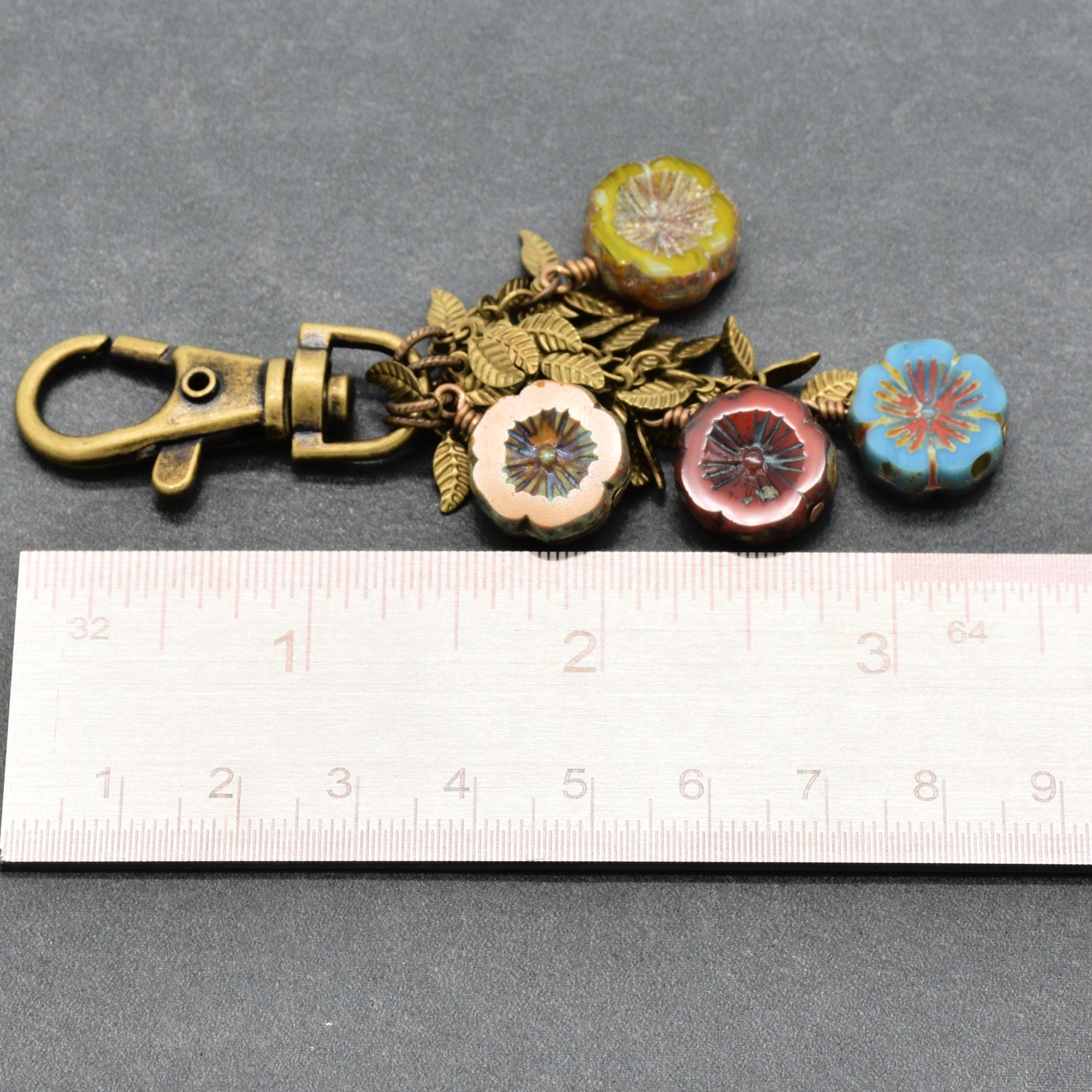 An antique bronze keychain with four flower charms in yellow, green, red, and blue, displayed next to a white ruler on a dark gray background - made with love in Colorado by Ruby Acorn.