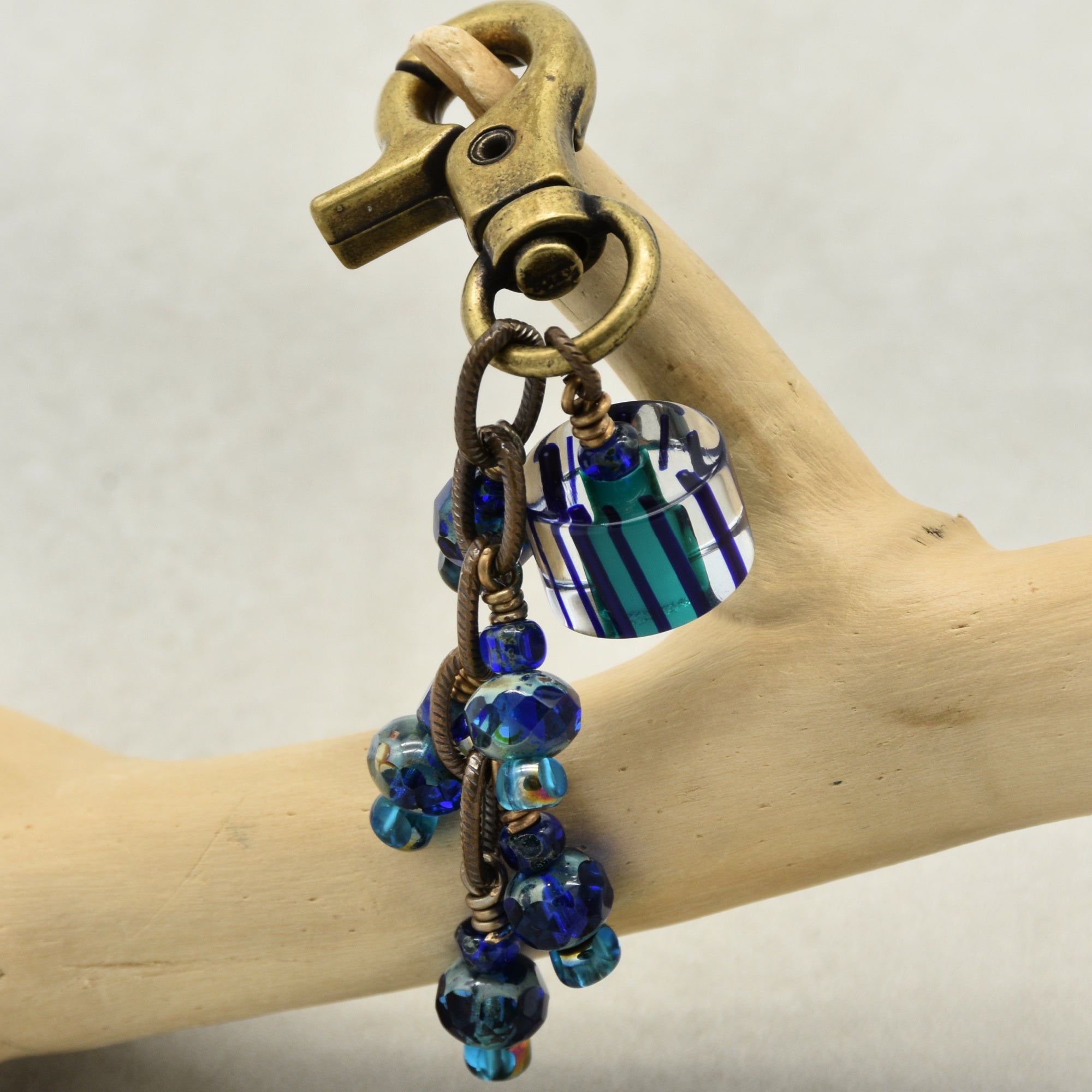 Brass keychain with blue beads and a striped cane  glass bead on a wooden branch.