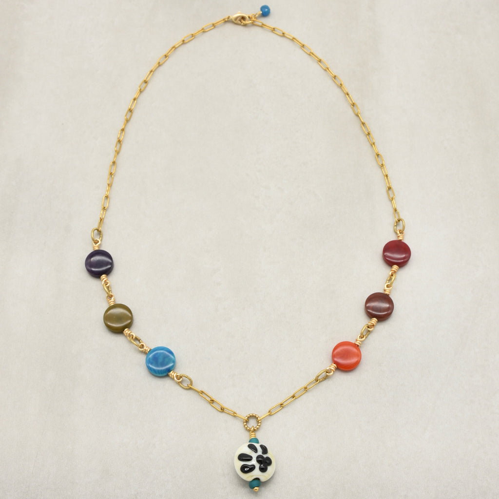 A gold chain necklace with colorful round beads in purple, olive green, light blue, orange-red, and burgundy, featuring a white paw print pendant  and small turquoise accents, displayed on a light beige textured background - made with love in Colorado by Ruby Acorn.