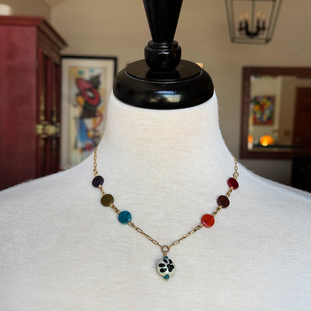 A beaded necklace displayed on a white fabric mannequin bust. The necklace features a gold-tone chain with multiple colorful round beads in dark purple, olive green, light blue, red, and orange, ending in a small black-and-white paw print pendant - made with love in Colorado by Ruby Acorn.