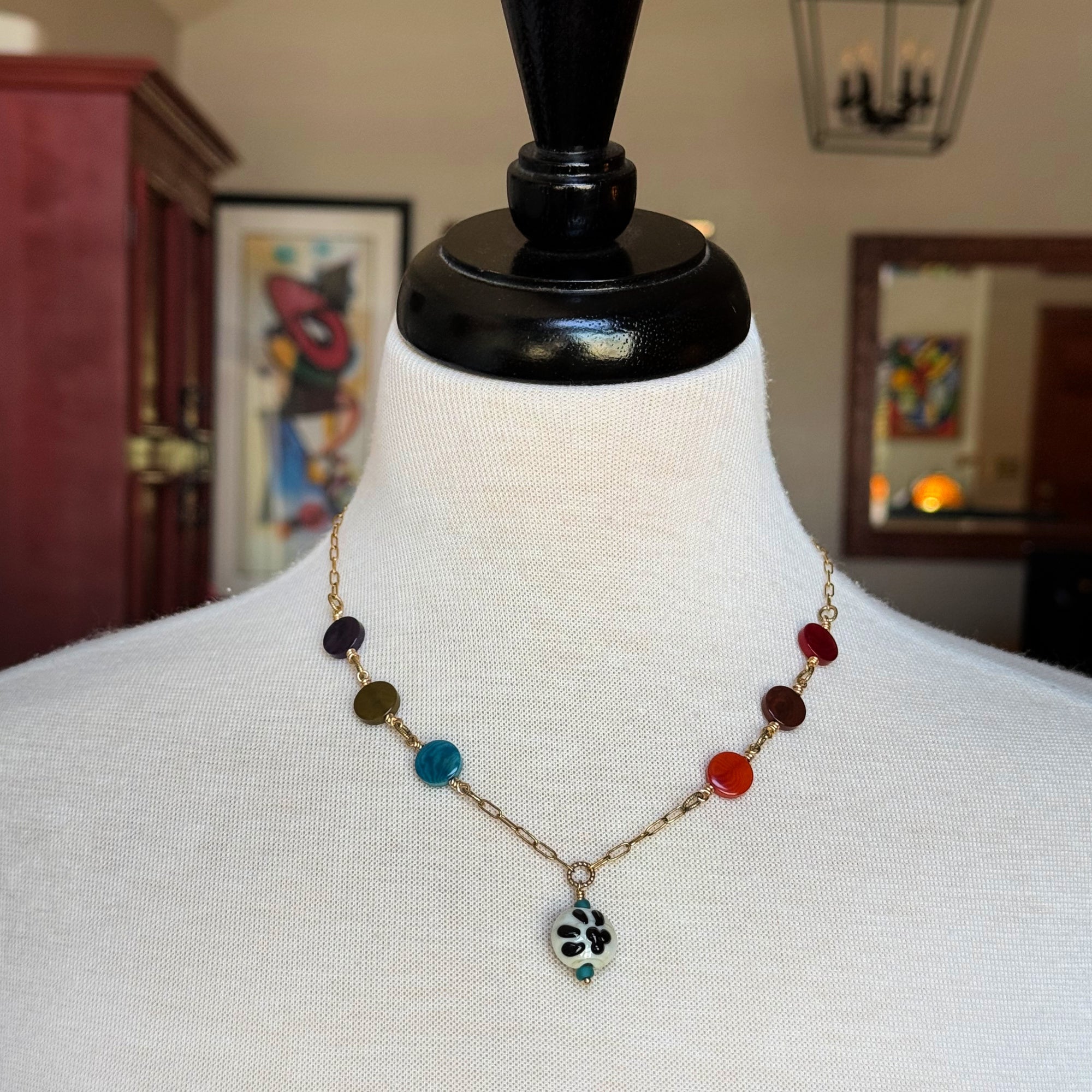 A beaded necklace displayed on a white fabric mannequin bust. The necklace features a gold-tone chain with multiple colorful round beads in dark purple, olive green, light blue, red, and orange, ending in a small black-and-white paw print pendant - made with love in Colorado by Ruby Acorn.