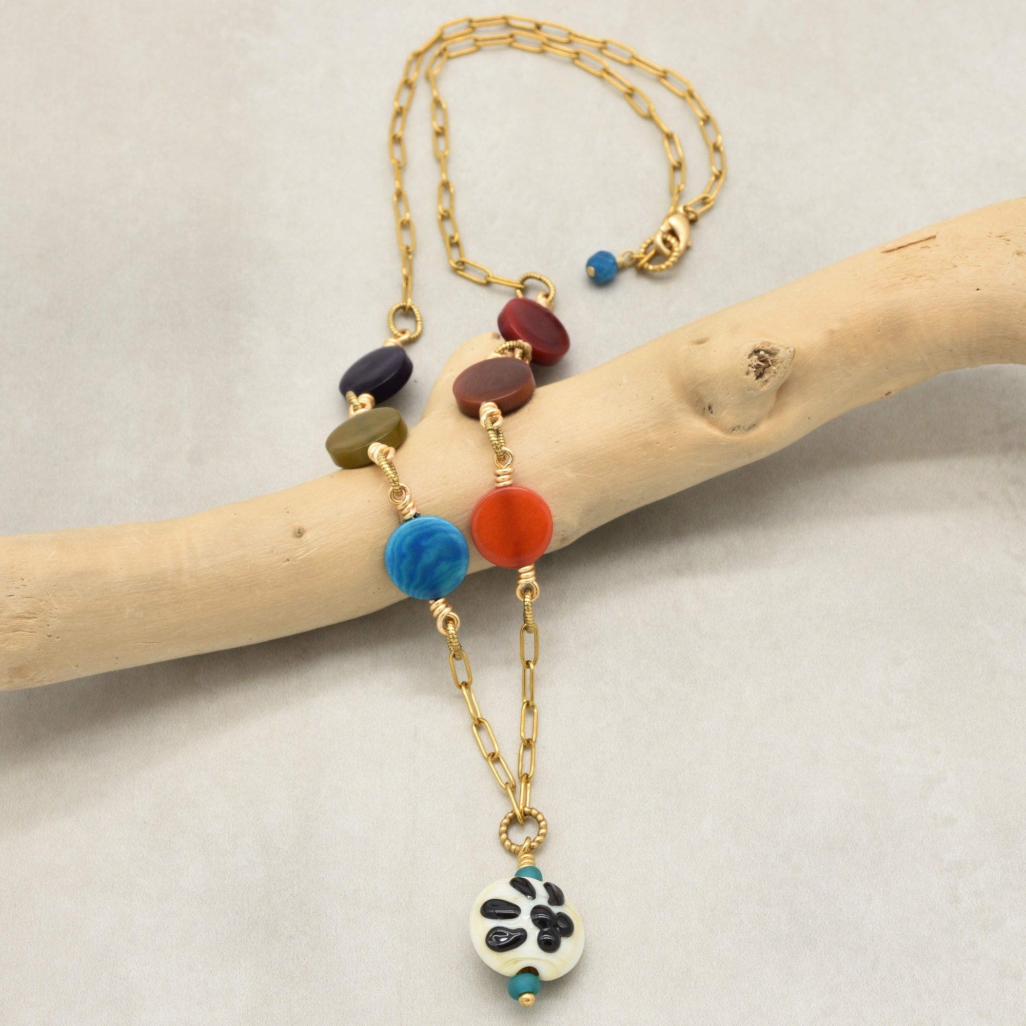 A bohemian-style gold chain necklace with colorful round beads and a white pendant featuring black paw print, displayed on a light wood branch against a neutral background - made with love in Colorado by Ruby Acorn.