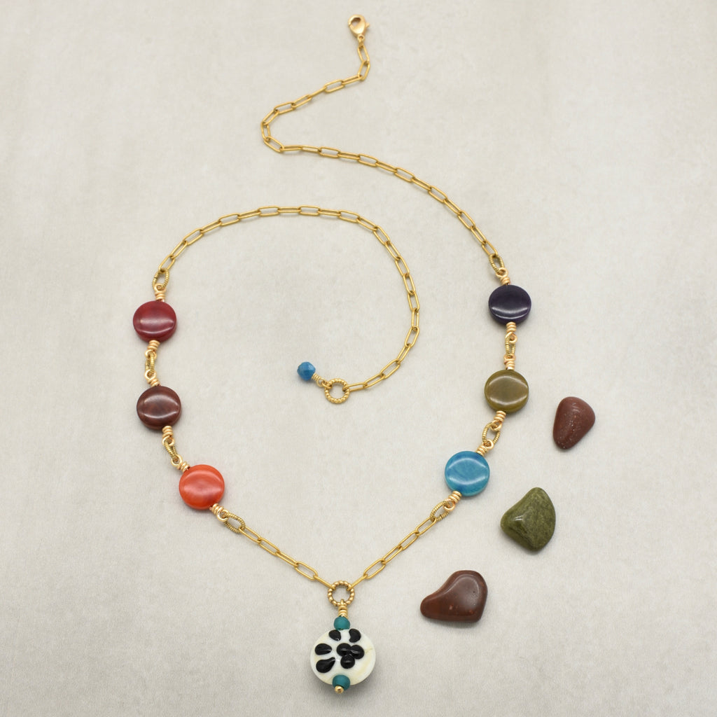A gold chain necklace with colorful polished stone beads and a white enamel paw print pendant, displayed on a light textured background with small loose stones nearby - made with love in Colorado by Ruby Acorn.