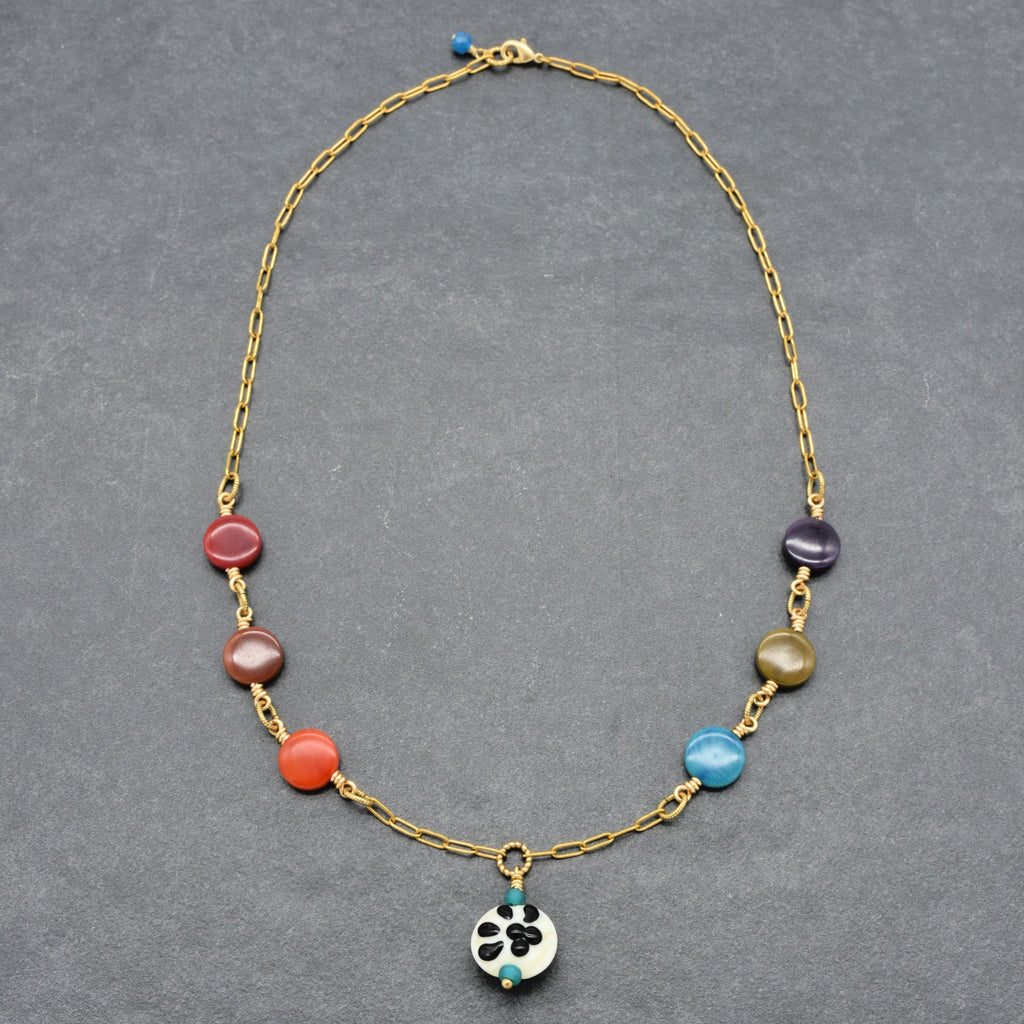A gold chain necklace featuring a series of colorful round beads in red, orange, purple, green, and blue, with a small white and black paw print focal at the center, displayed on a dark gray textured background - made with love in Colorado by Ruby Acorn.