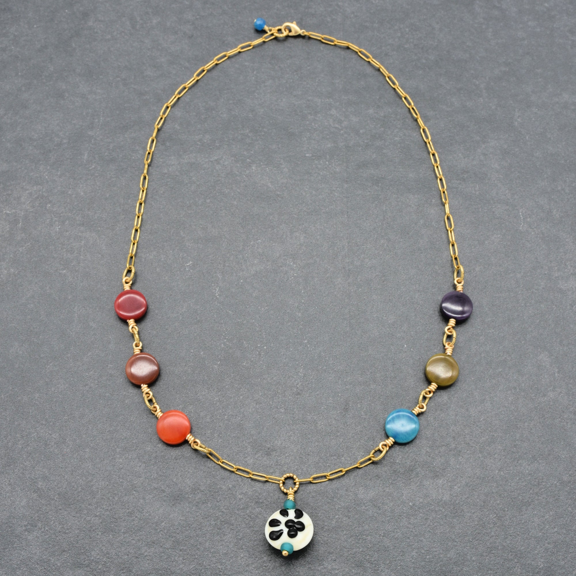 A gold chain necklace featuring a series of colorful round beads in red, orange, purple, green, and blue, with a small white and black paw print focal at the center, displayed on a dark gray textured background - made with love in Colorado by Ruby Acorn.