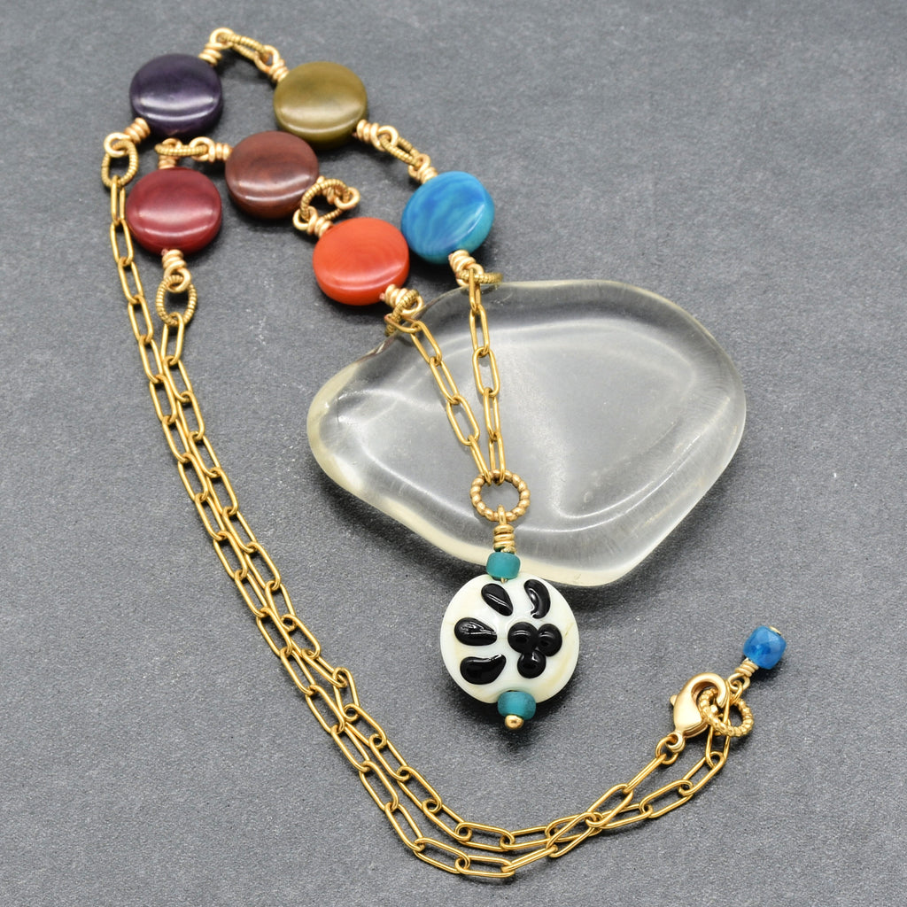 A gold-tone chain necklace with colorful round beads and a white pendant featuring black paw print, displayed on a gray textured background with a clear glass prop - made with love in Colorado by Ruby Acorn.