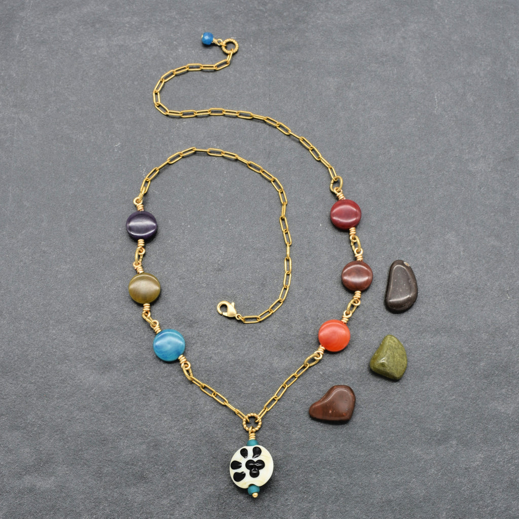 A gold chain necklace with colorful polished beads and a white circular pendant featuring a black paw print, displayed on a textured gray background with three loose accent stones nearby - made with love in Colorado by Ruby Acorn. 