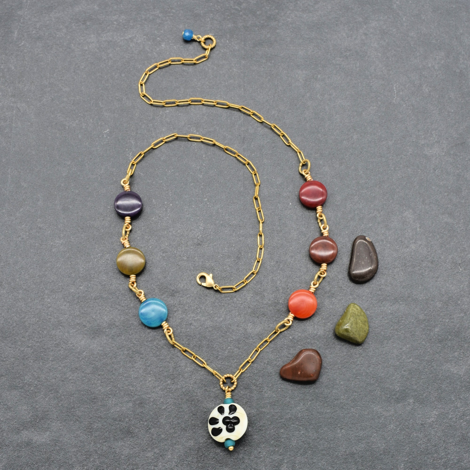 A gold chain necklace with colorful polished beads and a white circular pendant featuring a black paw print, displayed on a textured gray background with three loose accent stones nearby - made with love in Colorado by Ruby Acorn. 