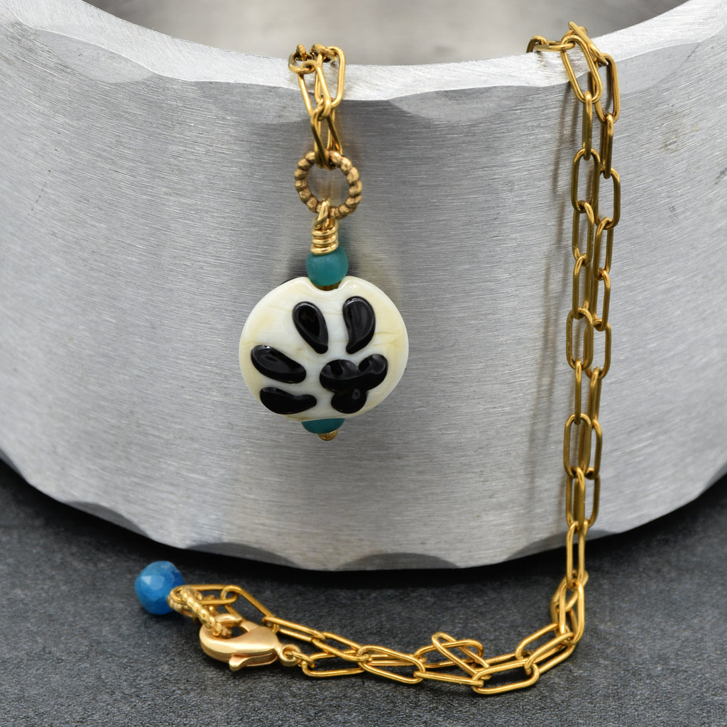 A gold chain necklace with a round white bead pendant featuring a black paw pattern and small turquoise accents, displayed on a textured gray background - made with love in Colorado by Ruby Acorn.