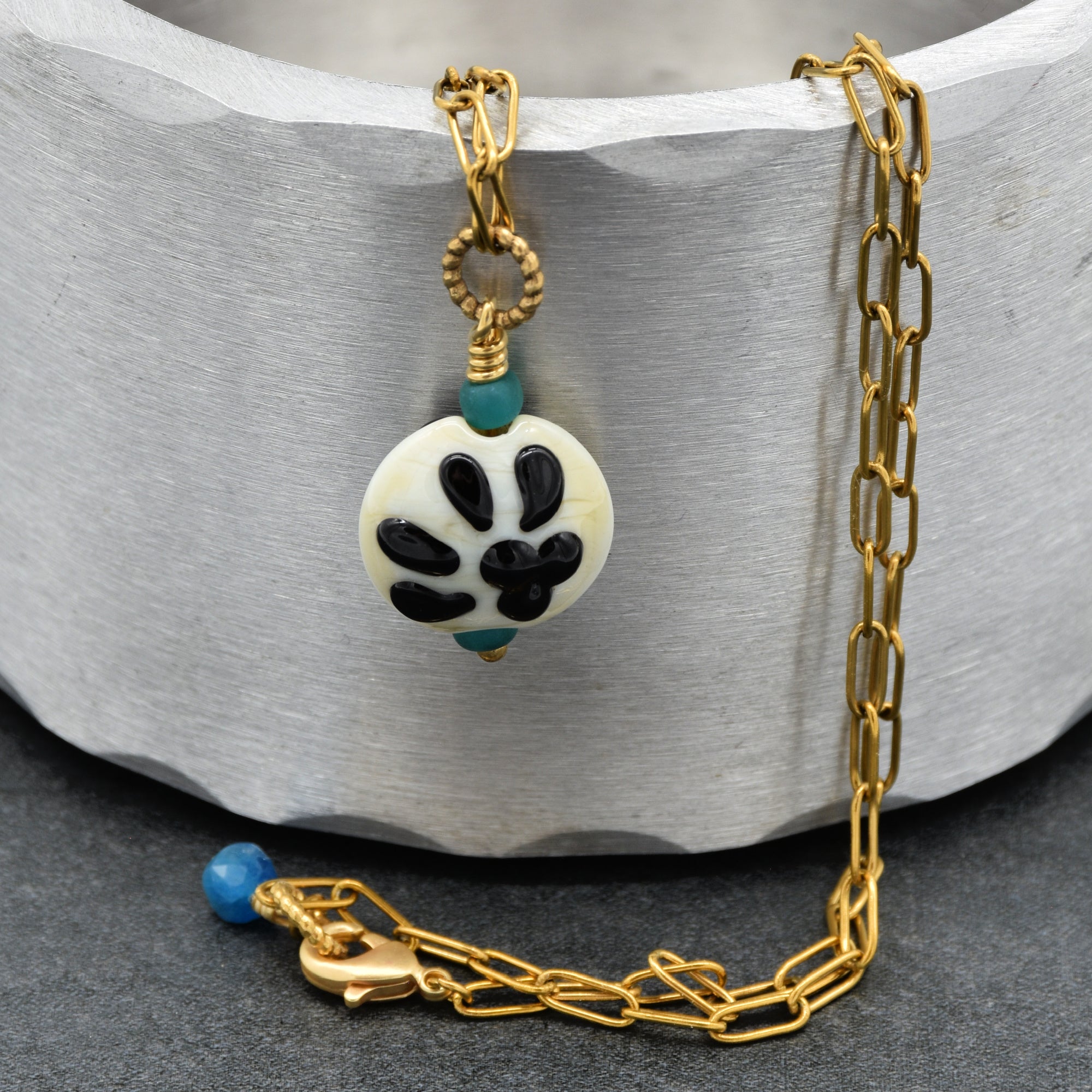 A gold chain necklace with a round white bead pendant featuring a black paw pattern and small turquoise accents, displayed on a textured gray background - made with love in Colorado by Ruby Acorn.