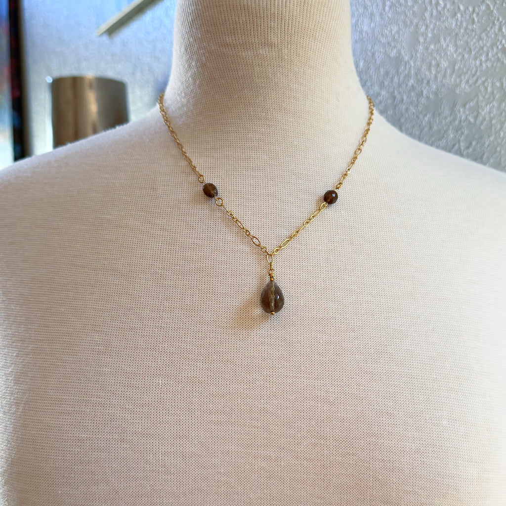 The image shows a necklace. It features a gold chain with small brown beads and a larger brown teardrop-shaped pendant hanging in the center.