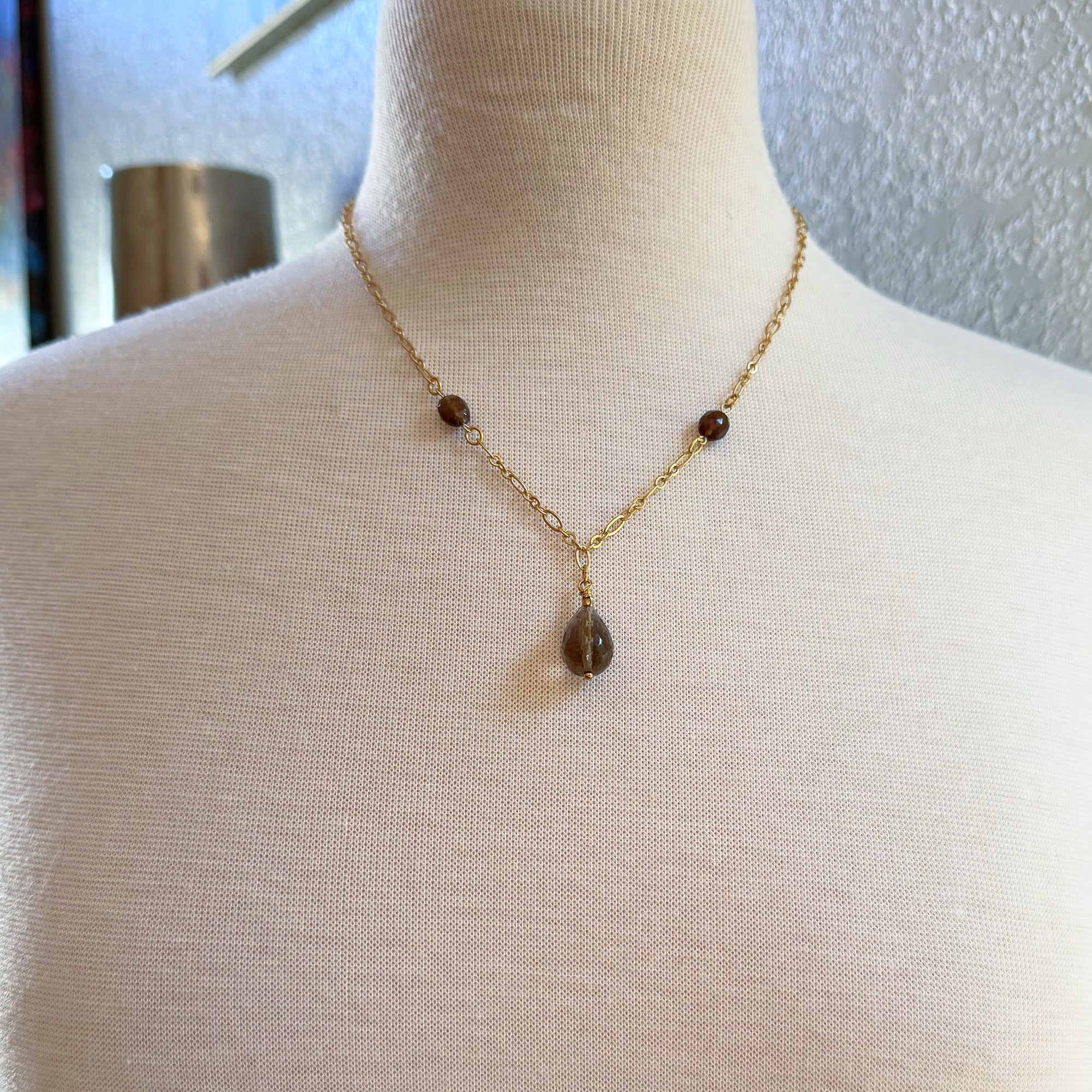 The image shows a necklace. It features a gold chain with small brown beads and a larger brown teardrop-shaped pendant hanging in the center.