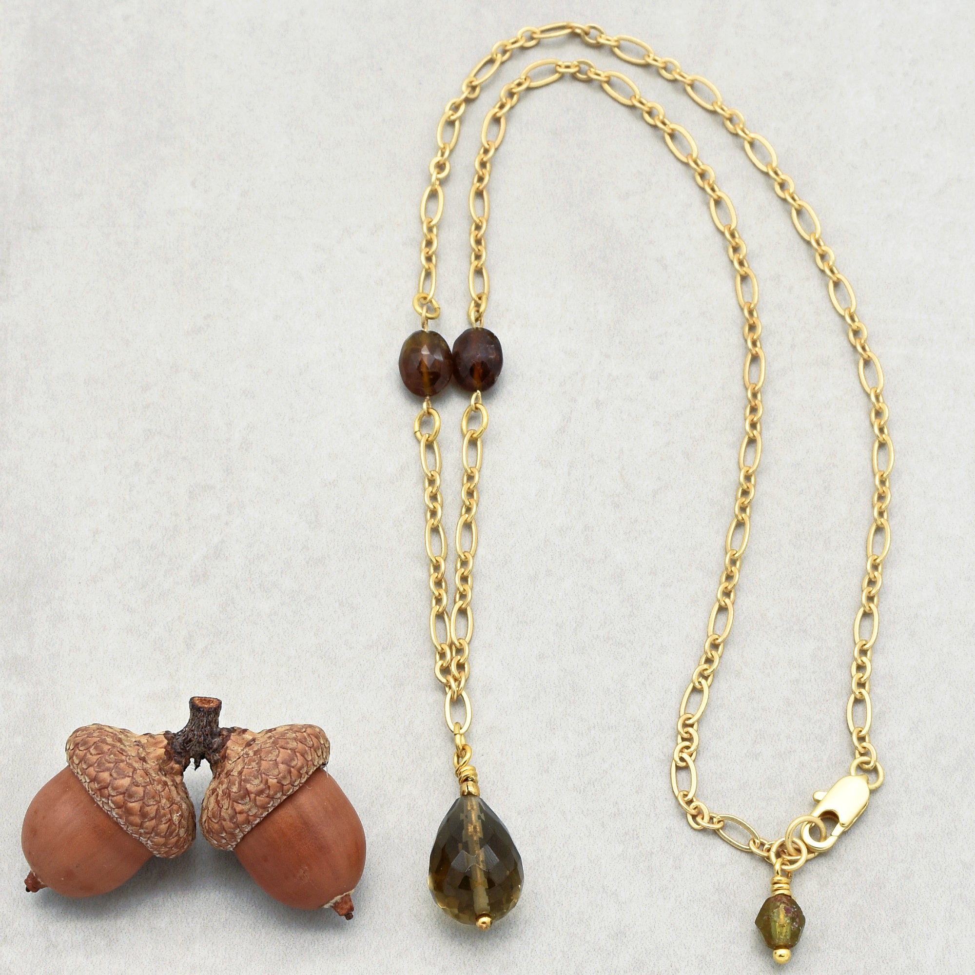 The image shows a necklace. It has a gold chain with two dark beads near the center and a larger, teardrop-shaped dark pendant hanging from it. There is also a small bead near the clasp. The necklace is displayed next to two acorns.