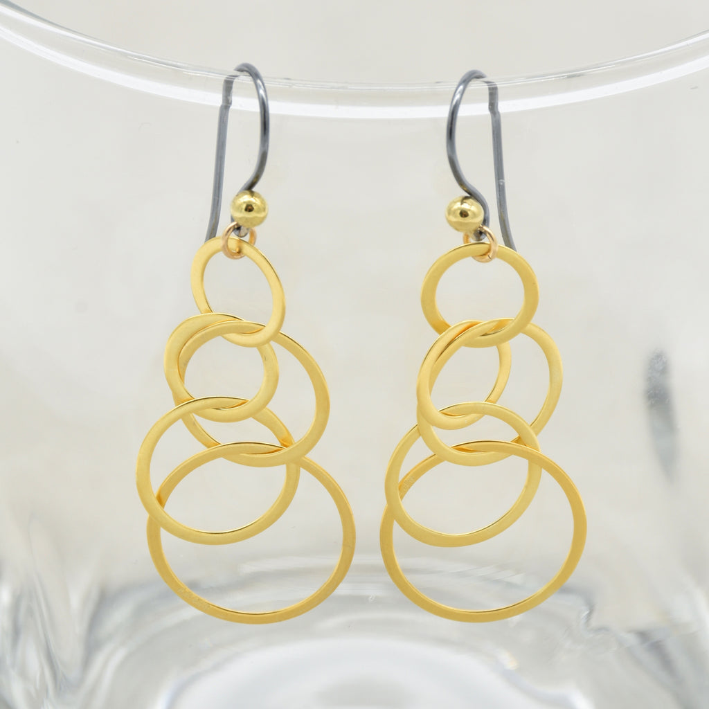 A pair of gold drop earrings with a stacked, interlocking circular ring design, hanging from black silver ear wires with small gold beads at the top - made with love by Ruby Acorn