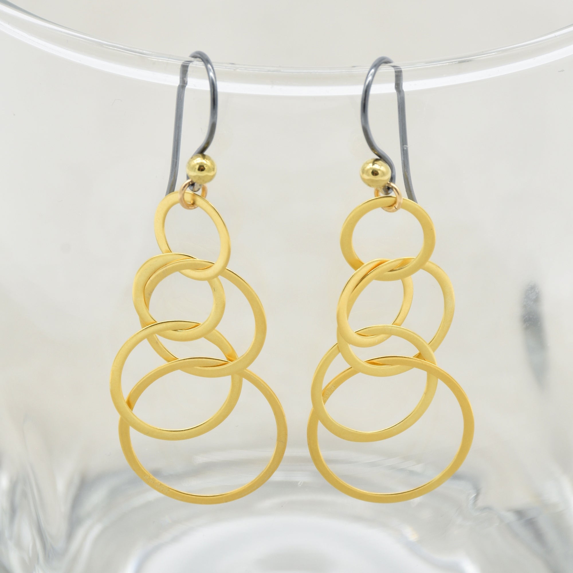 A pair of gold drop earrings with a stacked, interlocking circular ring design, hanging from black silver ear wires with small gold beads at the top - made with love by Ruby Acorn