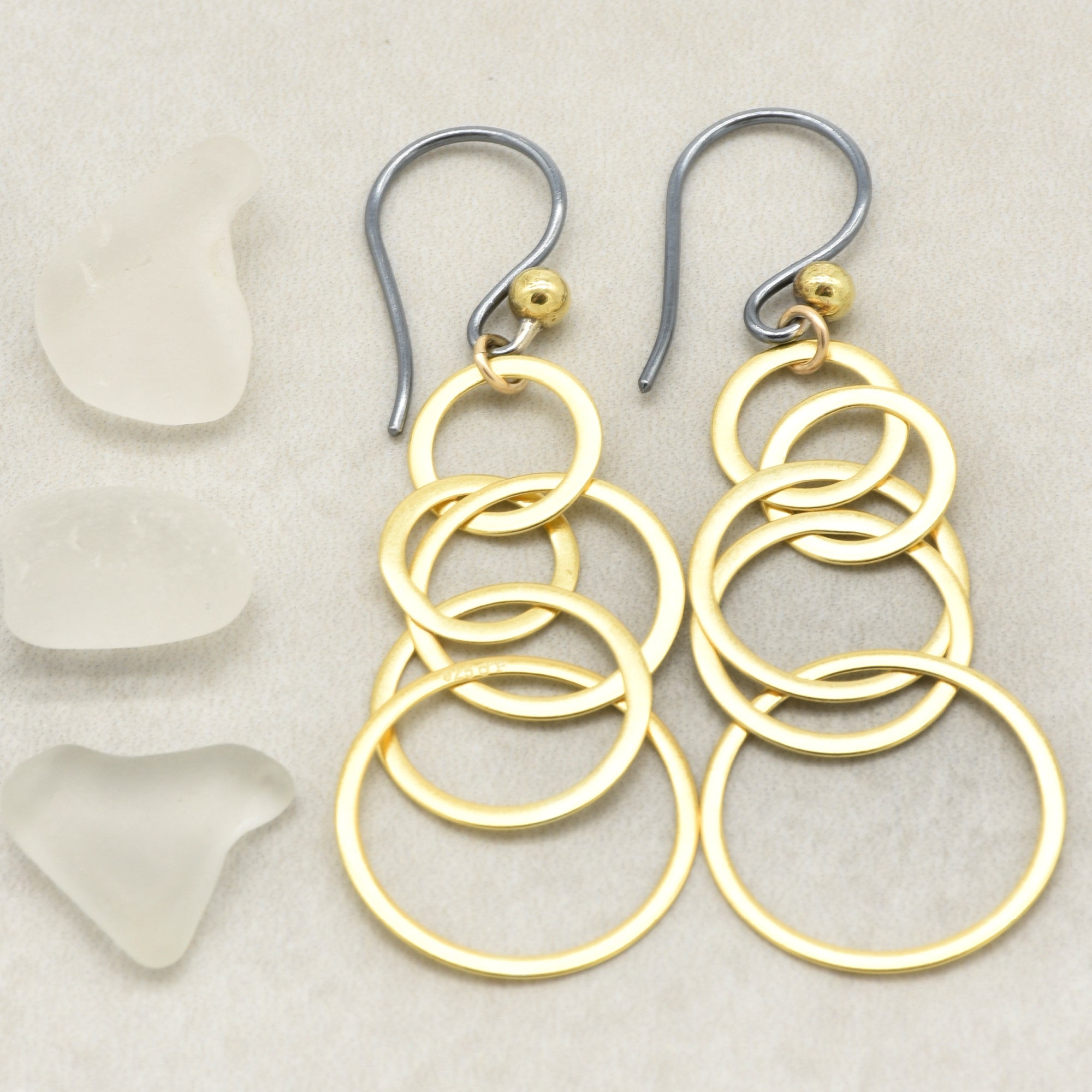 A pair of gold and silver drop earrings with interlocking circular rings, displayed on a light beige textured background next to three pieces of white sea glass - made with love by Ruby Acorn.